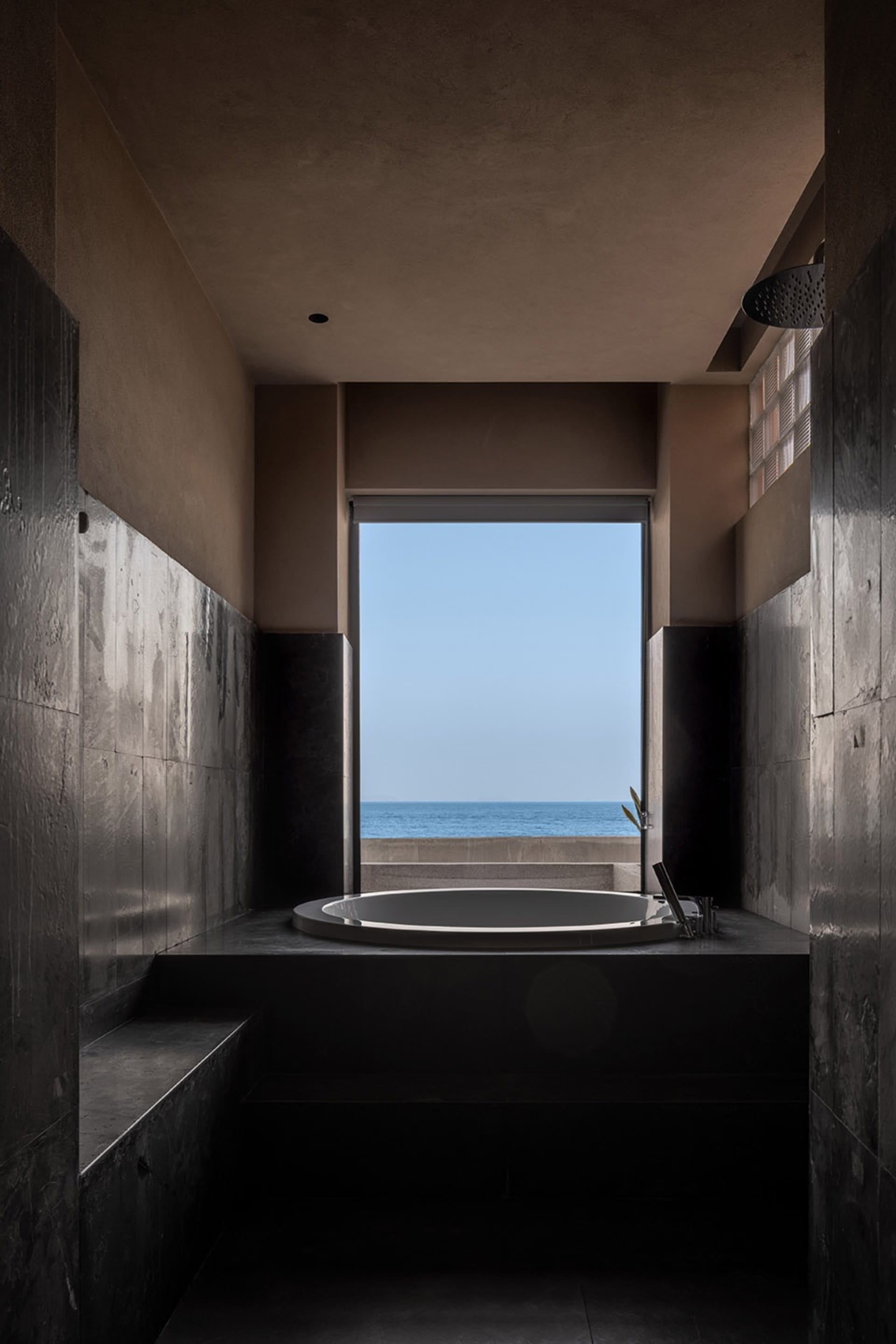 極簡浴室採用深色石材牆面與地板，配有圓形浴缸，可俯瞰海洋景色。