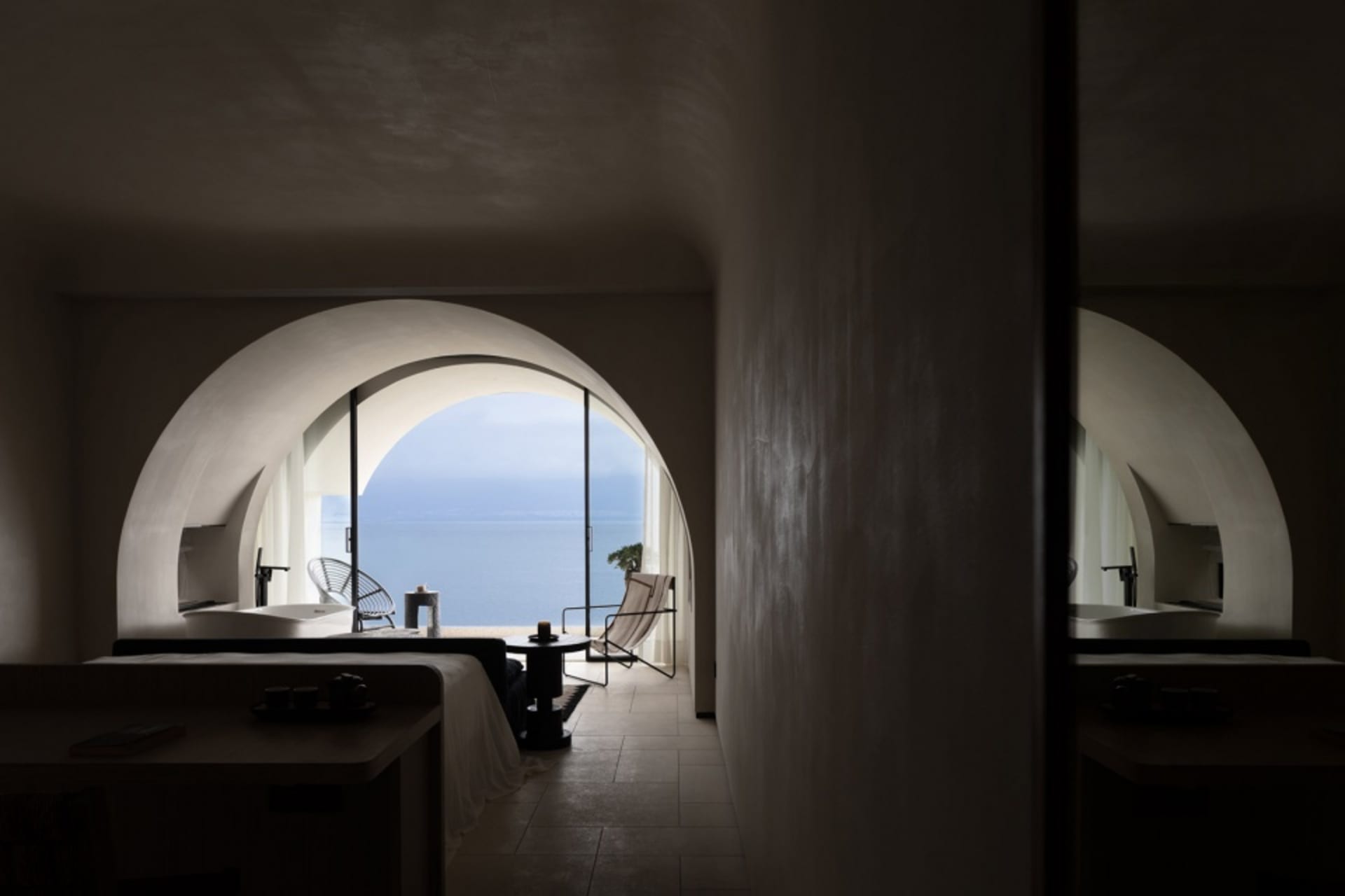 A minimalist bedroom featuring a bed in front of a large arched window overlooking the sea.