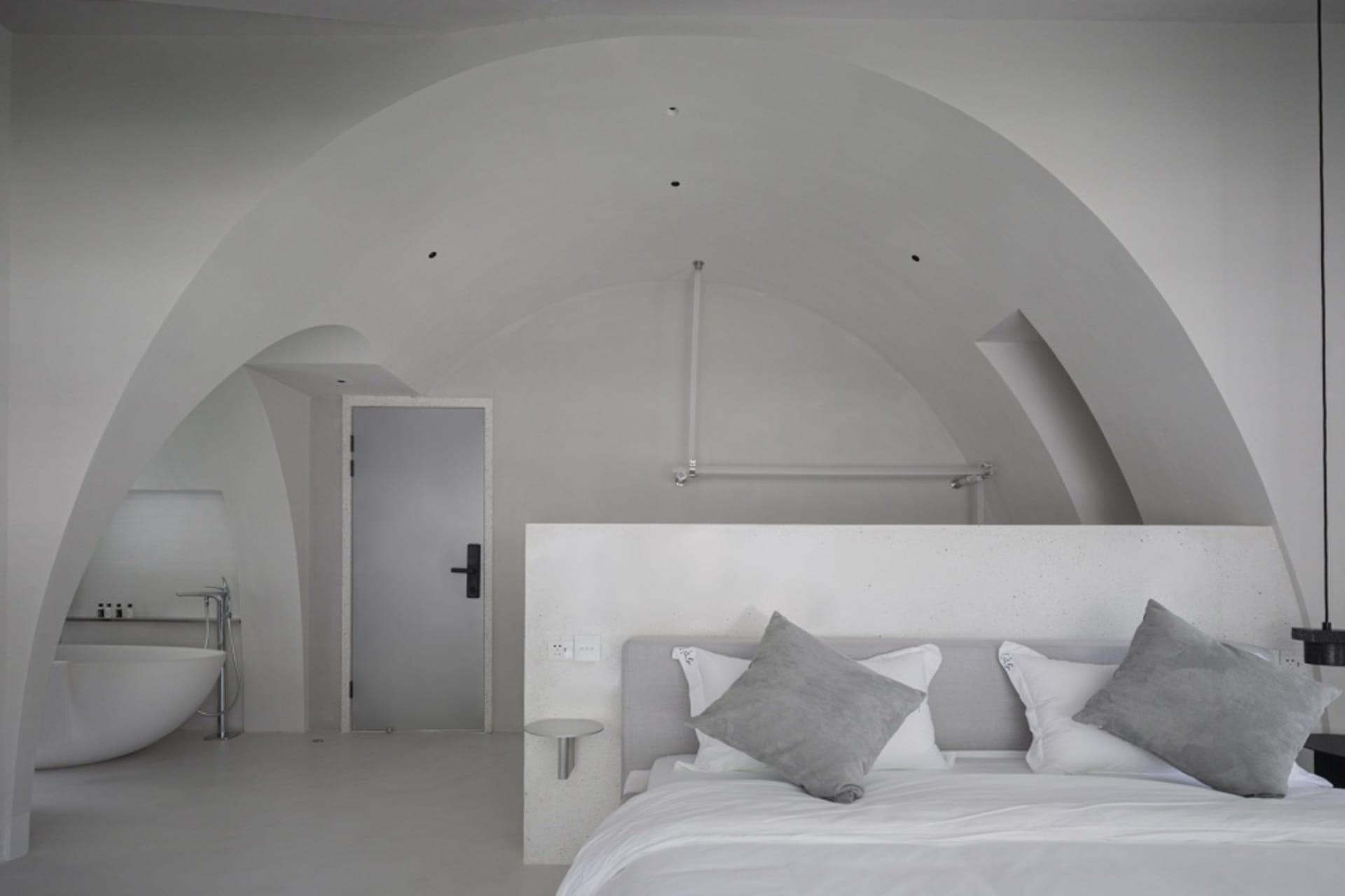 A minimalist bedroom with concrete walls, a white bed and pillows, and a sleek white bathtub in the corner.