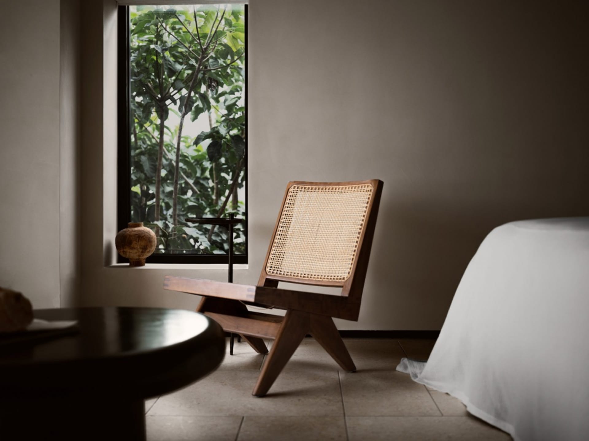 A minimalist bedroom with a wooden chair and table, natural light from a window, and a bed with a white sheet.