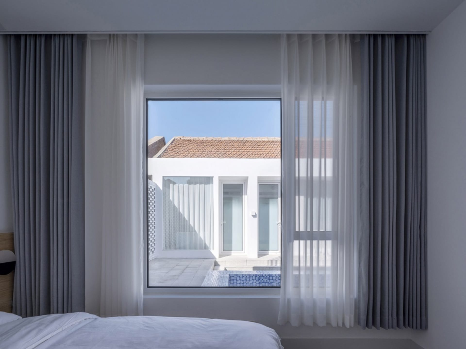 A modern residential bedroom featuring elegant curtains that let sunlight in, with a view of a white building with a tiled ro