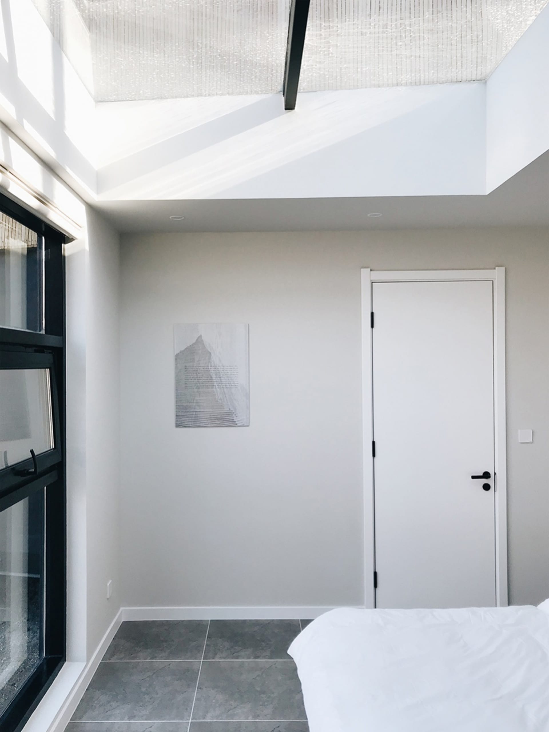 A minimalist bedroom featuring a concrete floor and walls, with a white bed and skylight.