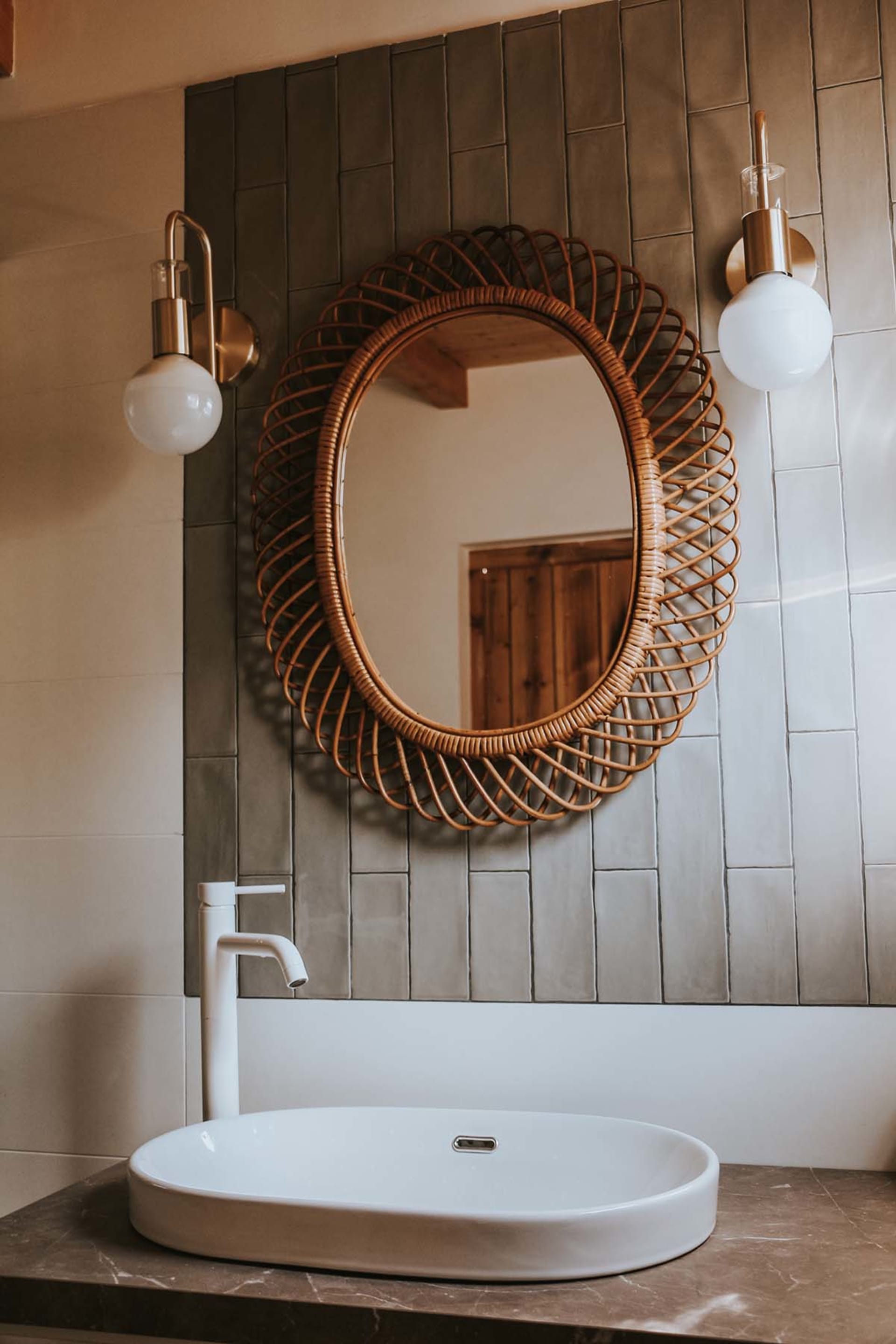 A modern bathroom with a tiled wall, oval sink, white faucet, and decorative mirror with a wicker frame.