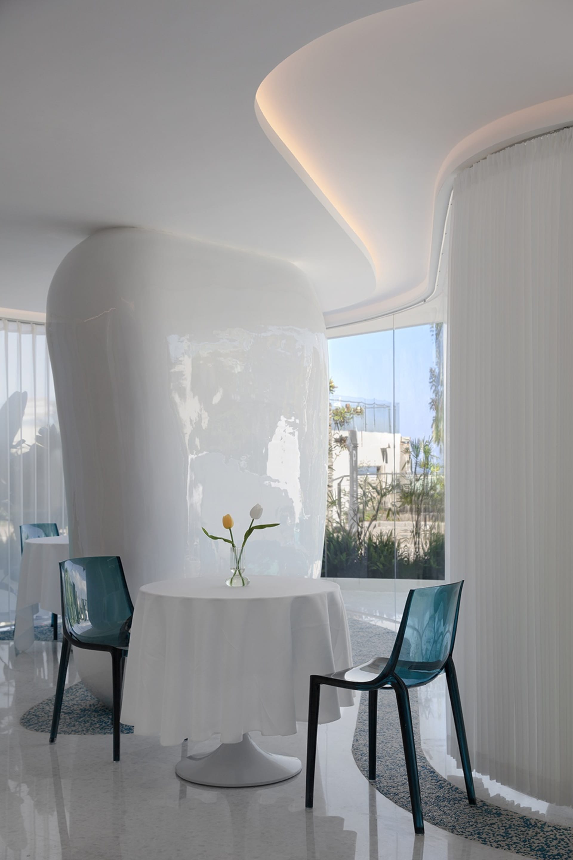 A minimalist restaurant interior with a white tablecloth, glass vase with tulips, and transparent blue plastic chairs.