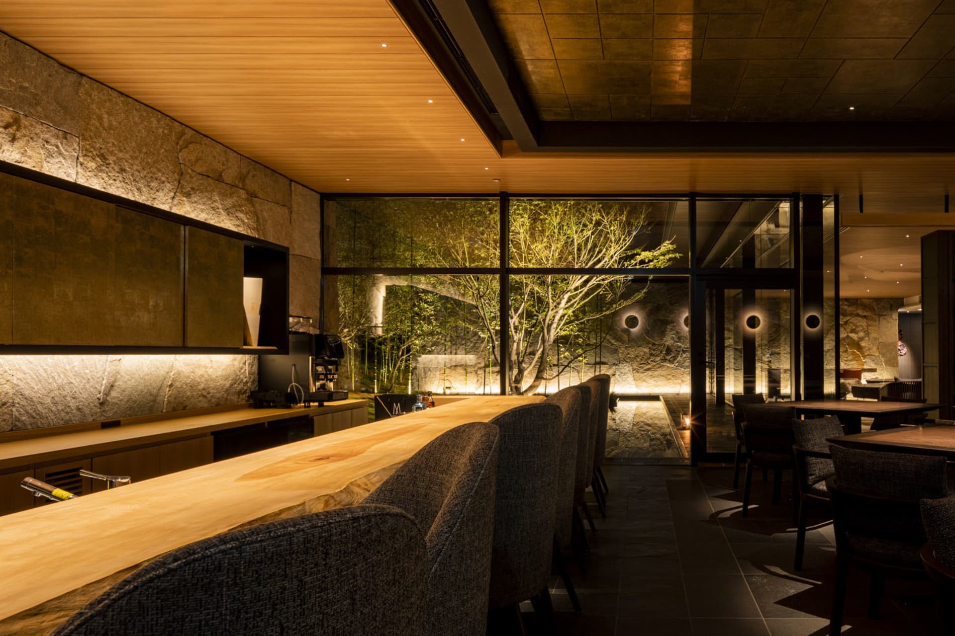 Industrial-style bar area with wooden counter, stone wall, and large windows showing vegetation outside.