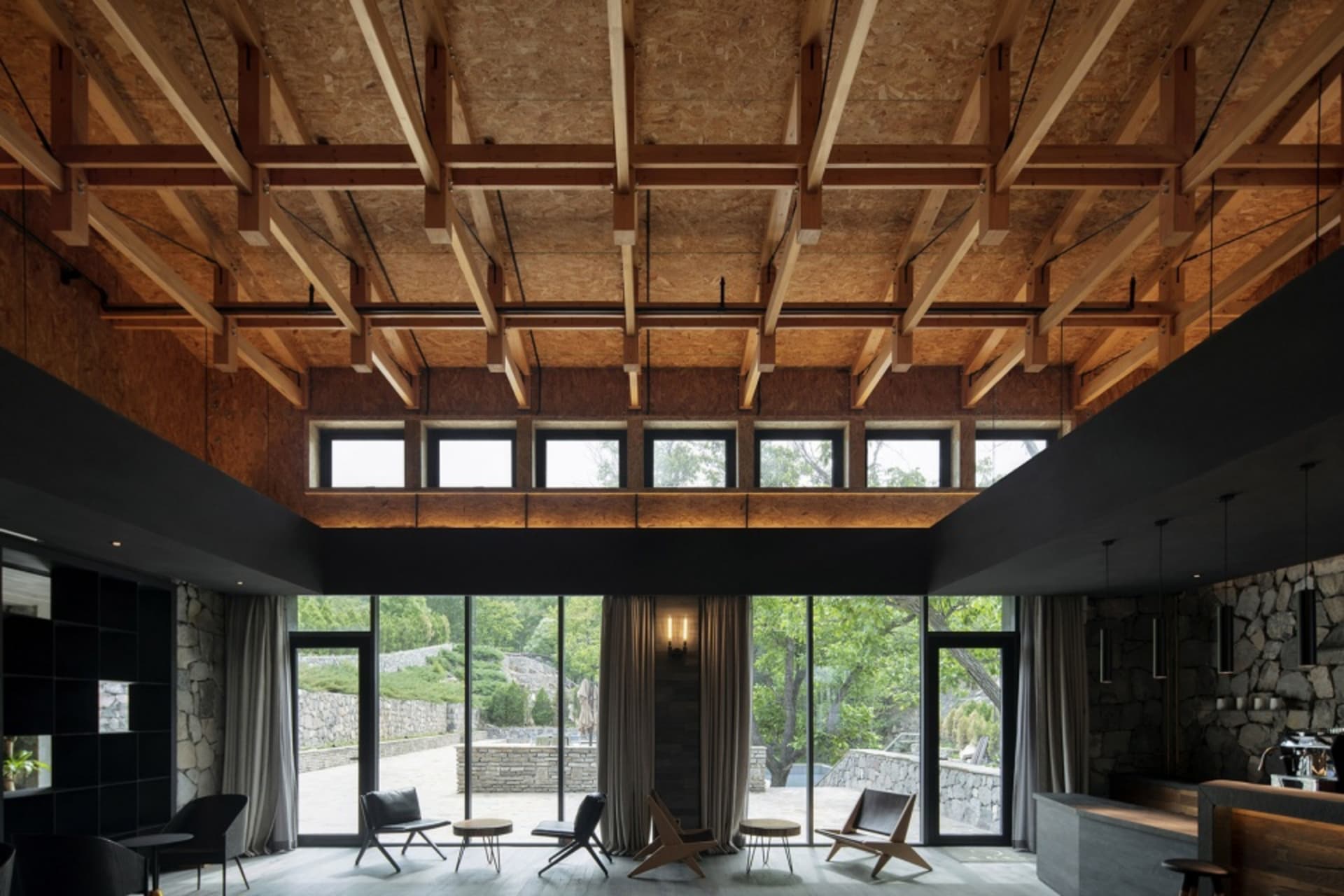 An industrial hotel suite featuring exposed wooden beams, brick walls, and a stone floor, with large windows offering a view