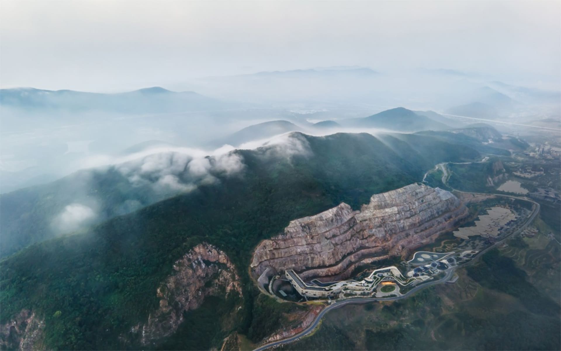 Aerial view of a mining site with layered rock formations and clouds near the mountains.
