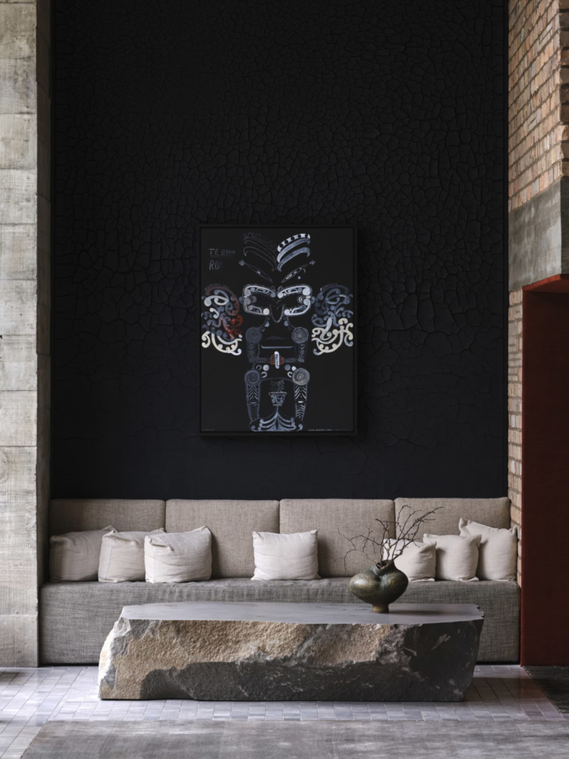 A modern living room features a concrete coffee table, fabric seating, and a black wall art piece with a tribal design.