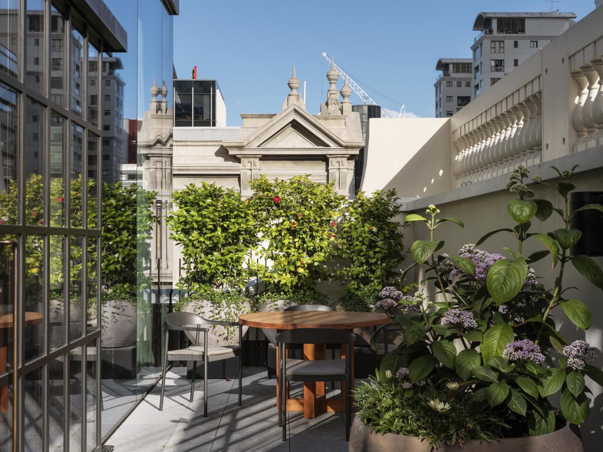 A modern rooftop garden with a wooden table, chairs, potted plants, and a view of neighboring buildings.