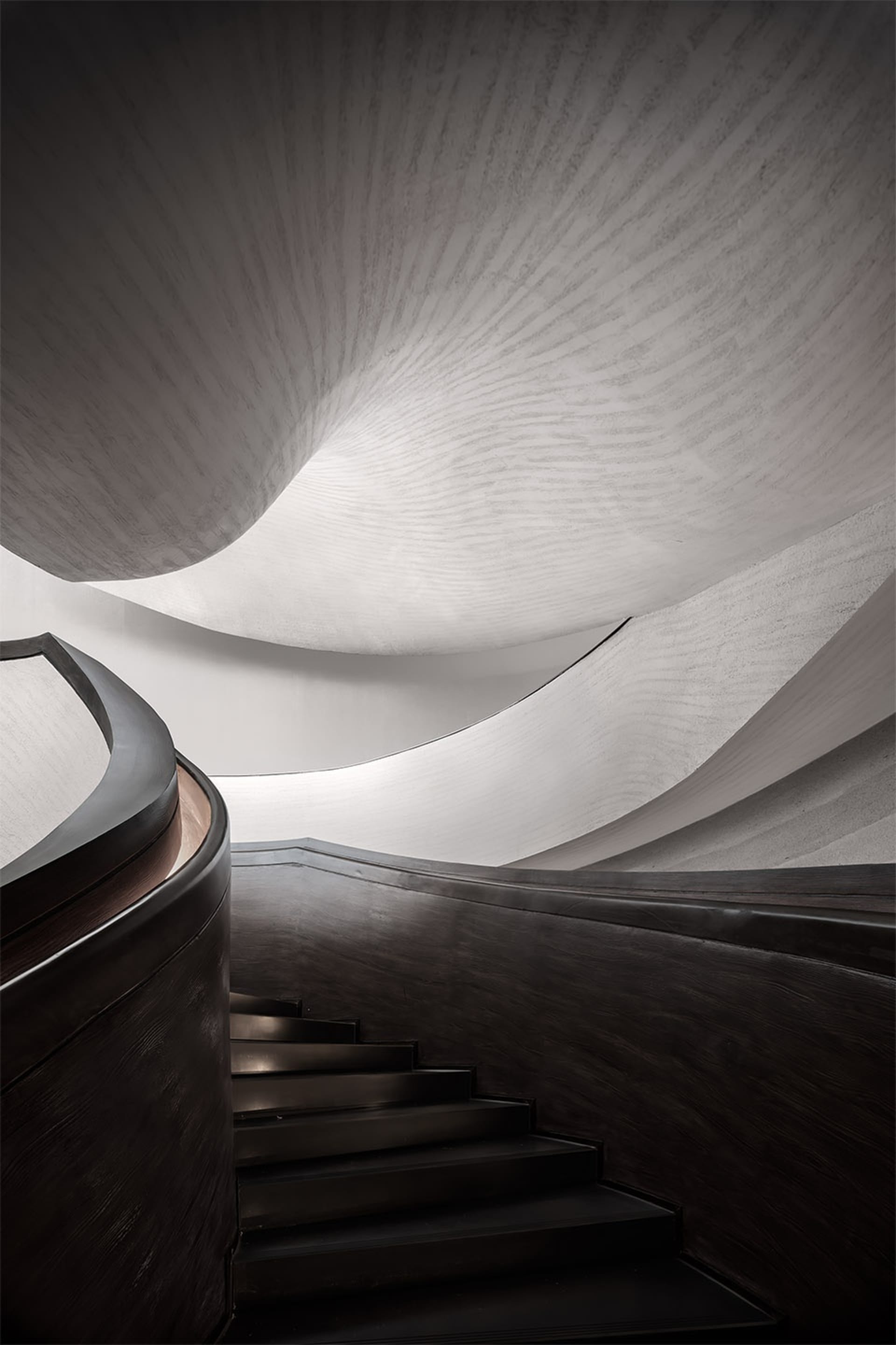 A sleek, modern staircase with curved metal steps and a dark wood railing, set against a textured, light-colored wall.