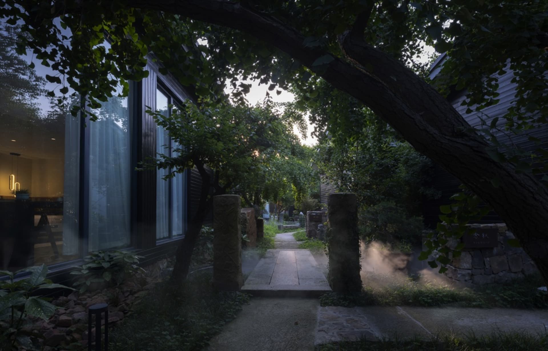 An evening view of a serene garden pathway with a stone path illuminated by mist and surrounded by lush greenery and trees.