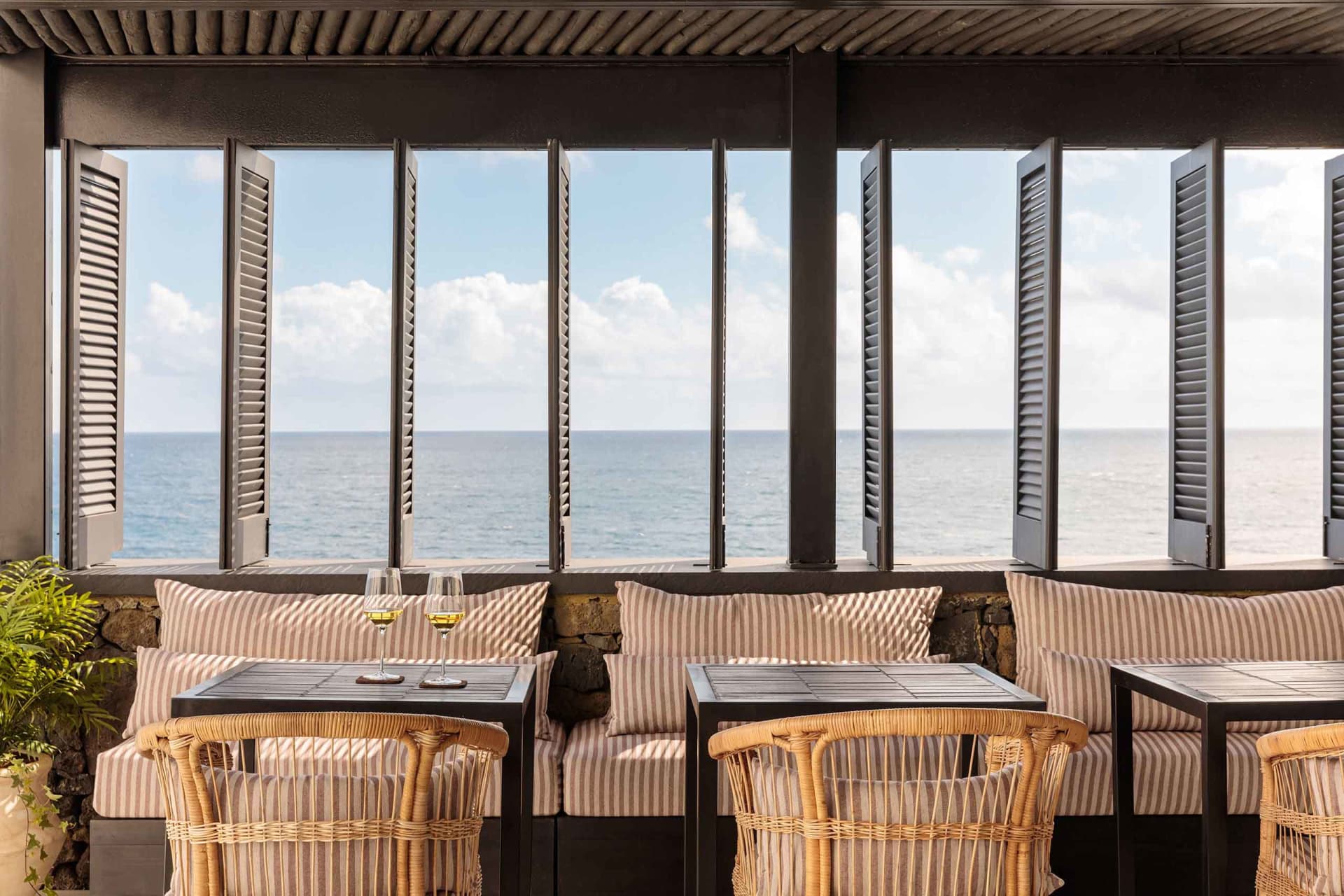 A coastal restaurant bar with a view of the ocean, featuring wooden furniture, stone accents, and curtains. Two wine glasses