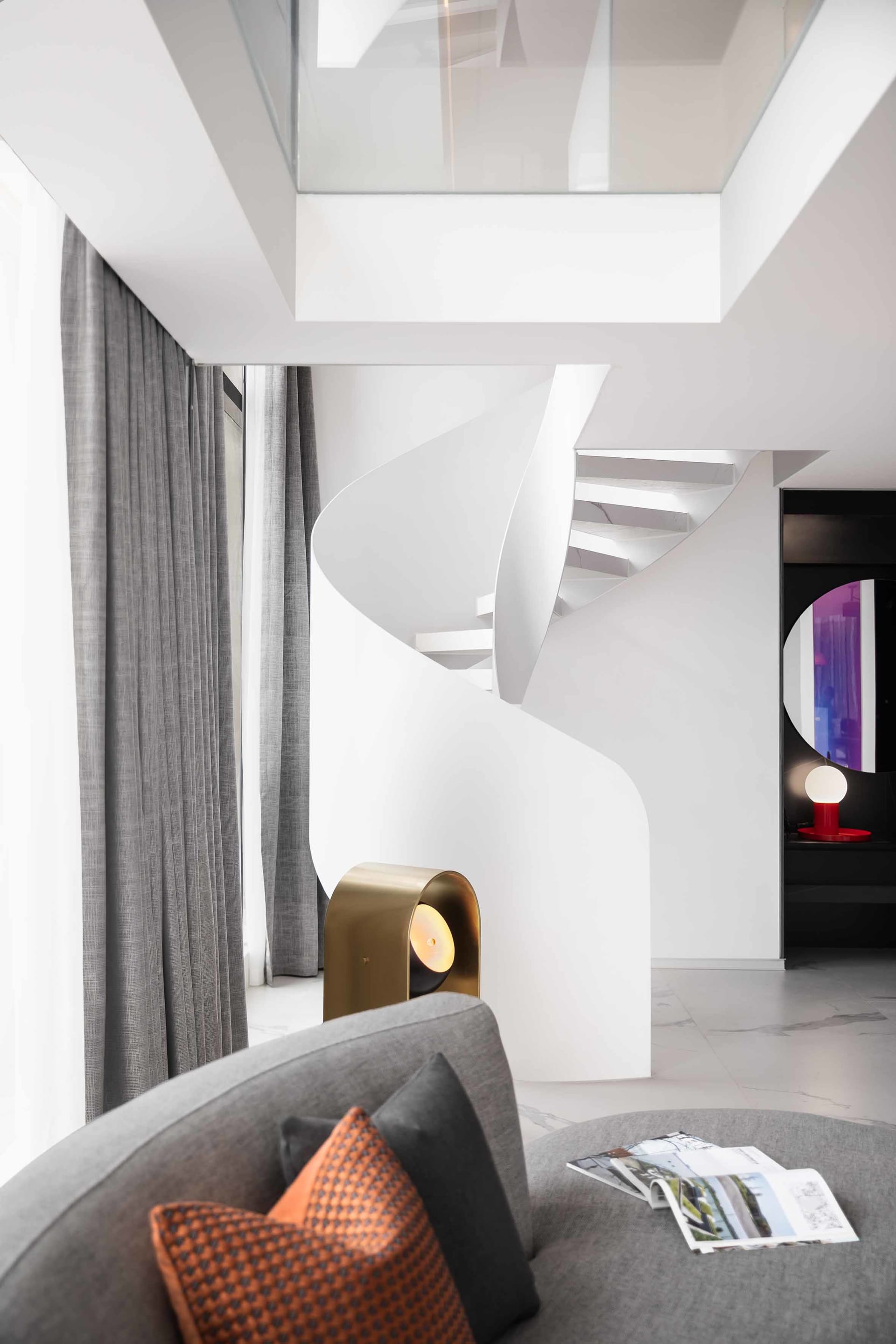 Modern living room with a metal and brass lamp, glass railing staircase, and grey fabric sofa with orange and black cushions.
