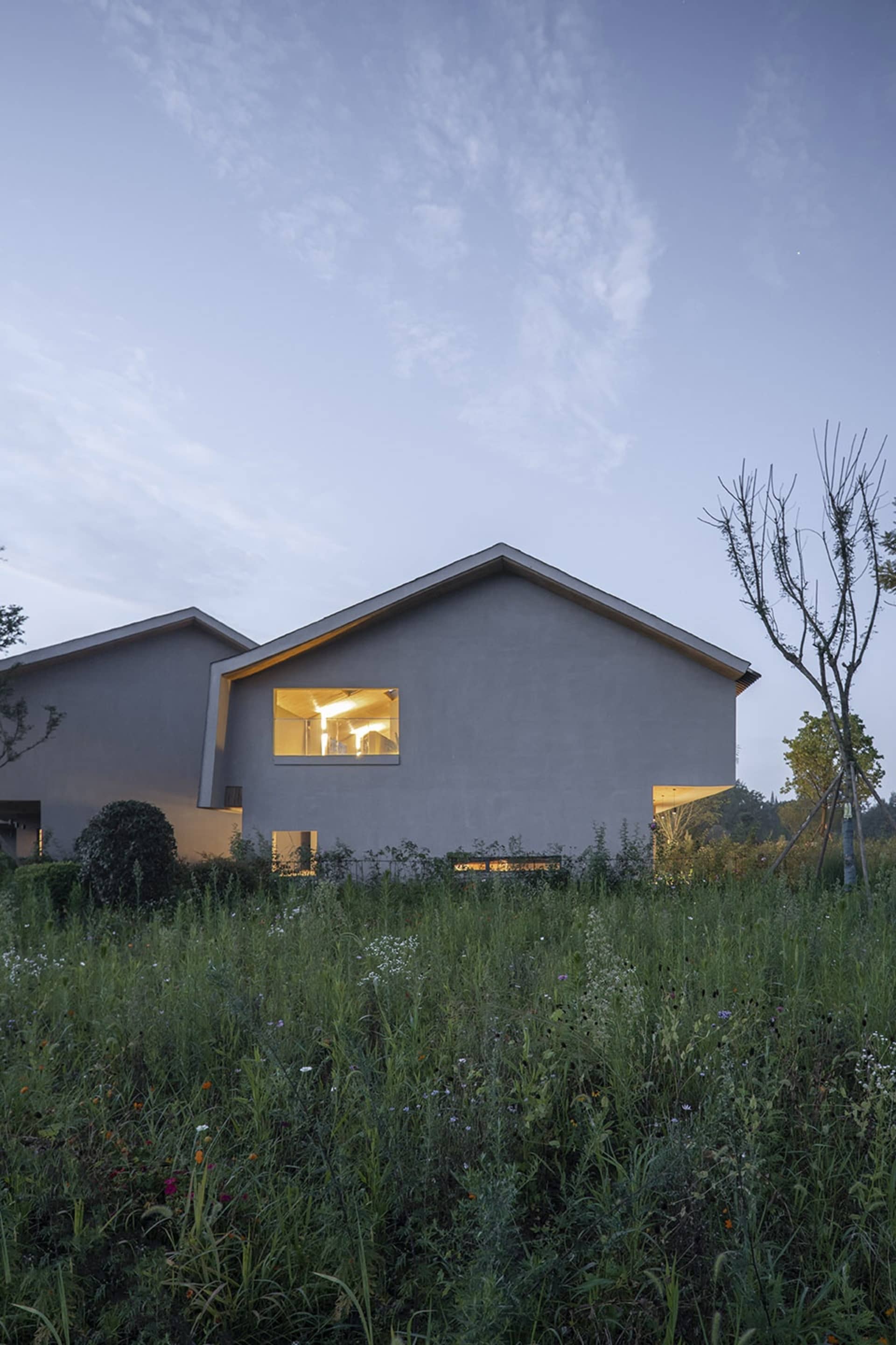 A minimalist residential concrete house with illuminated windows at dusk, surrounded by tall green grass