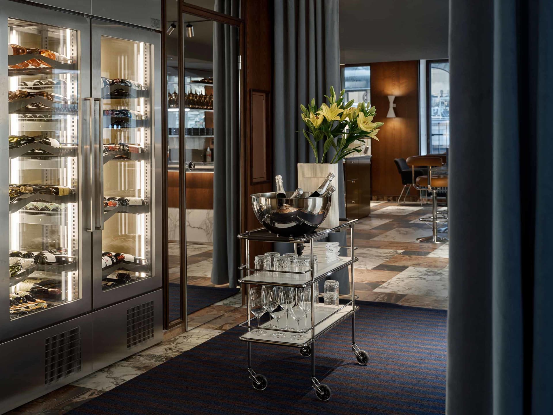 A metallic wine bar with wine bottles lined up in a glass case and a rolling cart filled with glasses and champagne bottles.