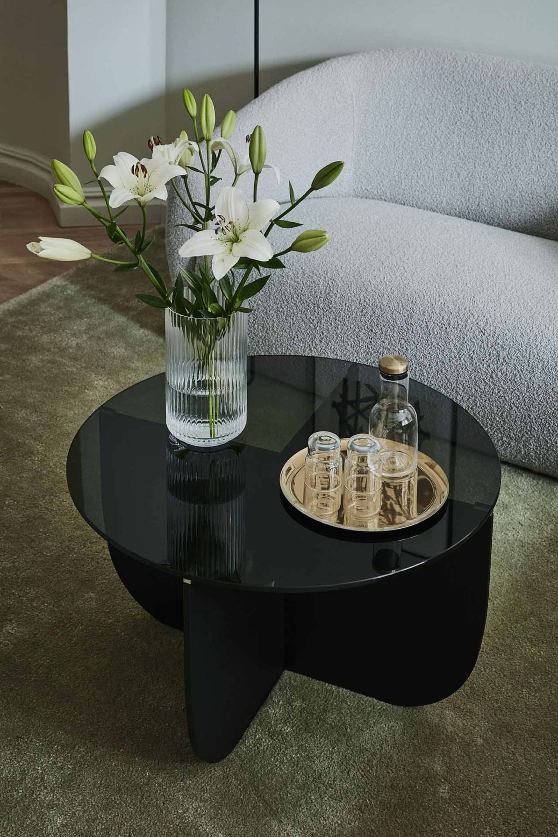 A modern interior design featuring a black glass side table with a gold tray, flowers in a vase, and a grey couch in the back
