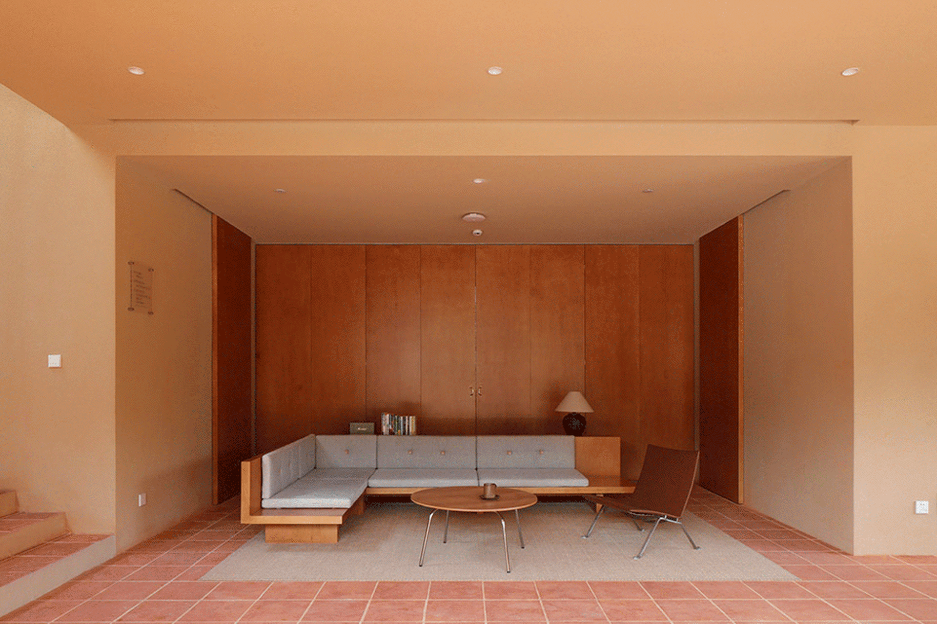 A modern, minimalist living room featuring a wooden bench-style sofa with a white cushion, a round wooden coffee table, and a