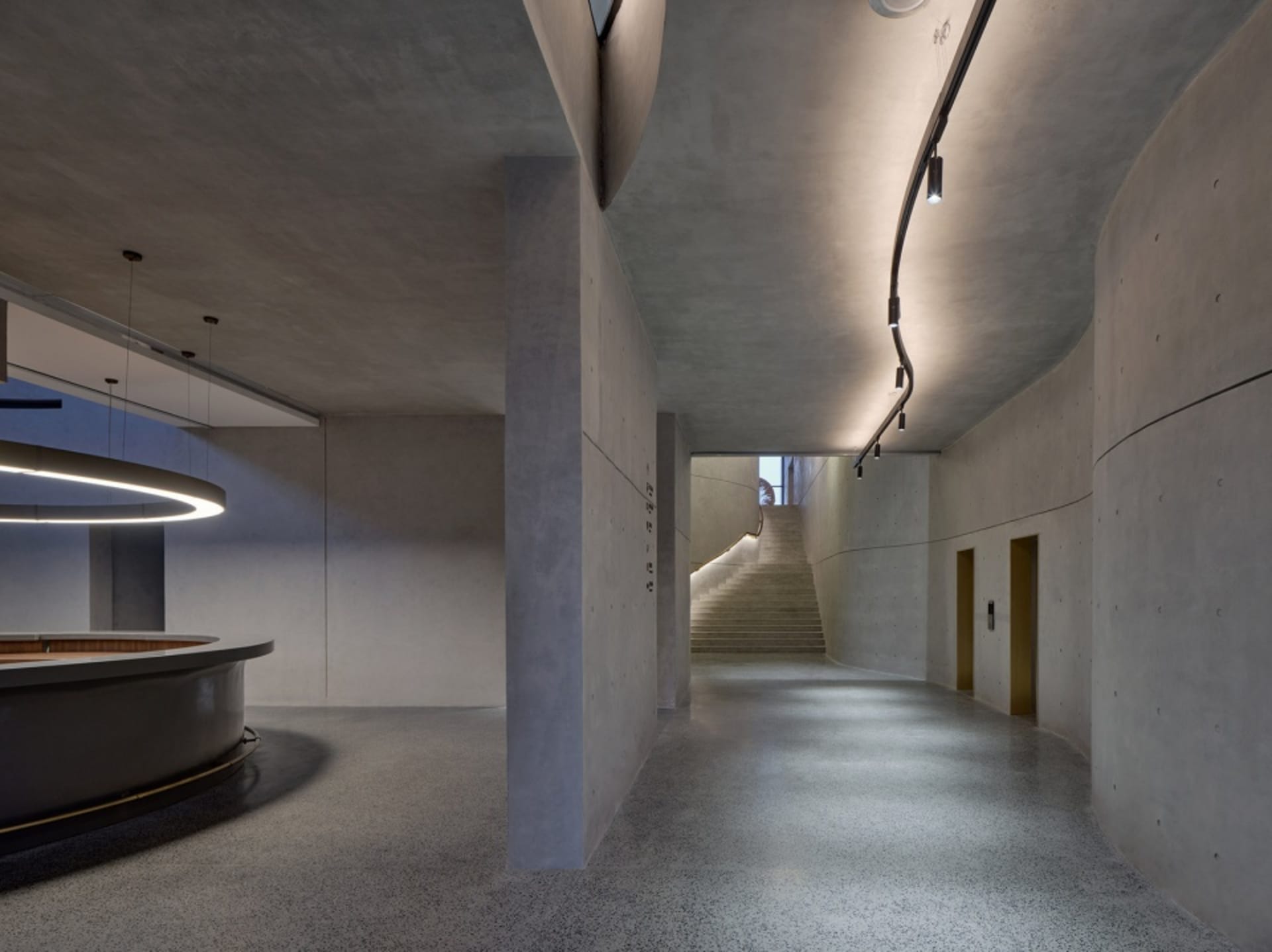 A modern office space with concrete walls and floors, featuring a curved reception desk and a staircase leading to another le