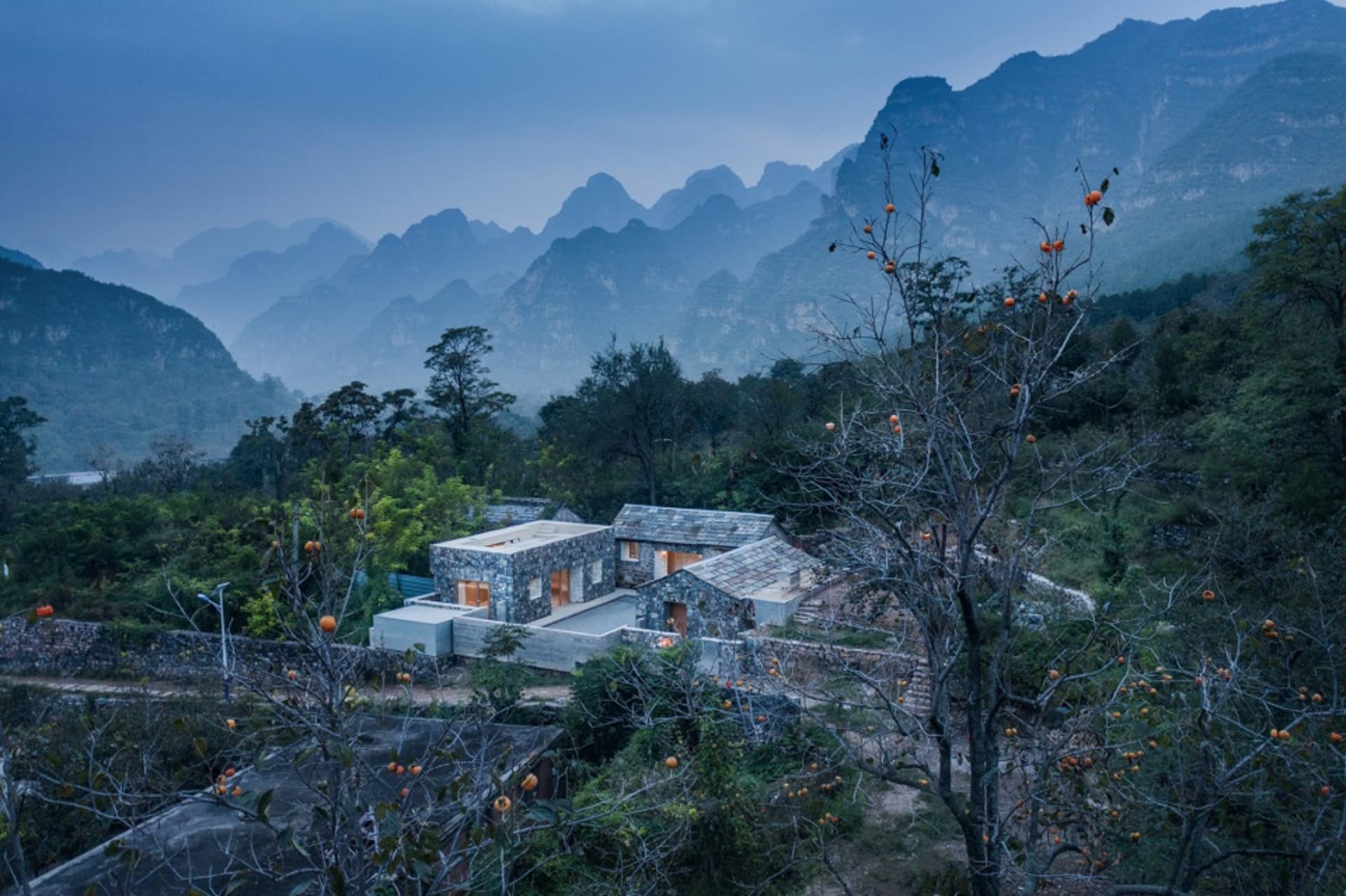 A modern residential building constructed from stone and concrete with wooden accents surrounded by lush greenery and mountai