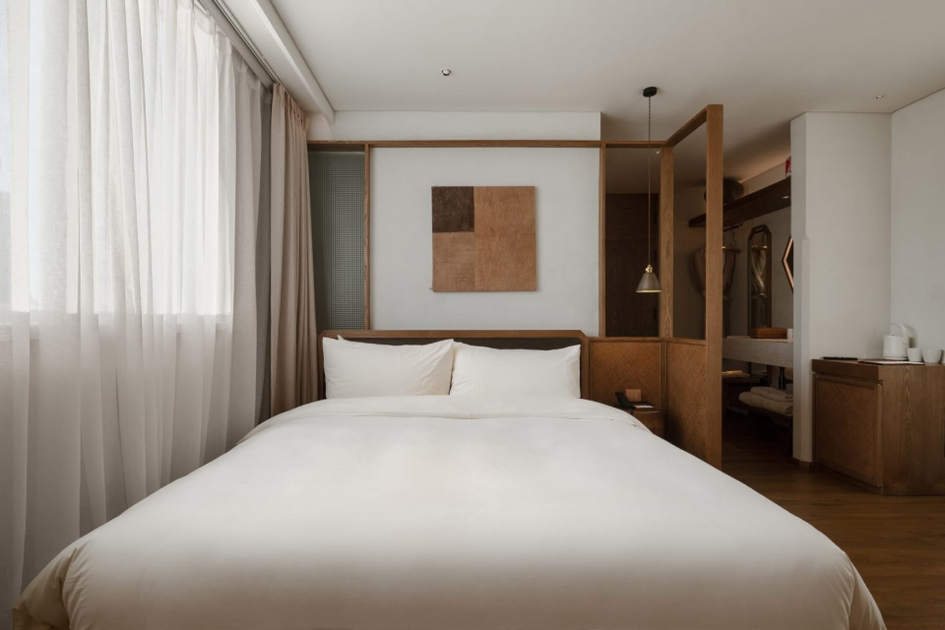 A modern minimalist bedroom with a large bed featuring white fabric, surrounded by wooden elements and a large window with sh