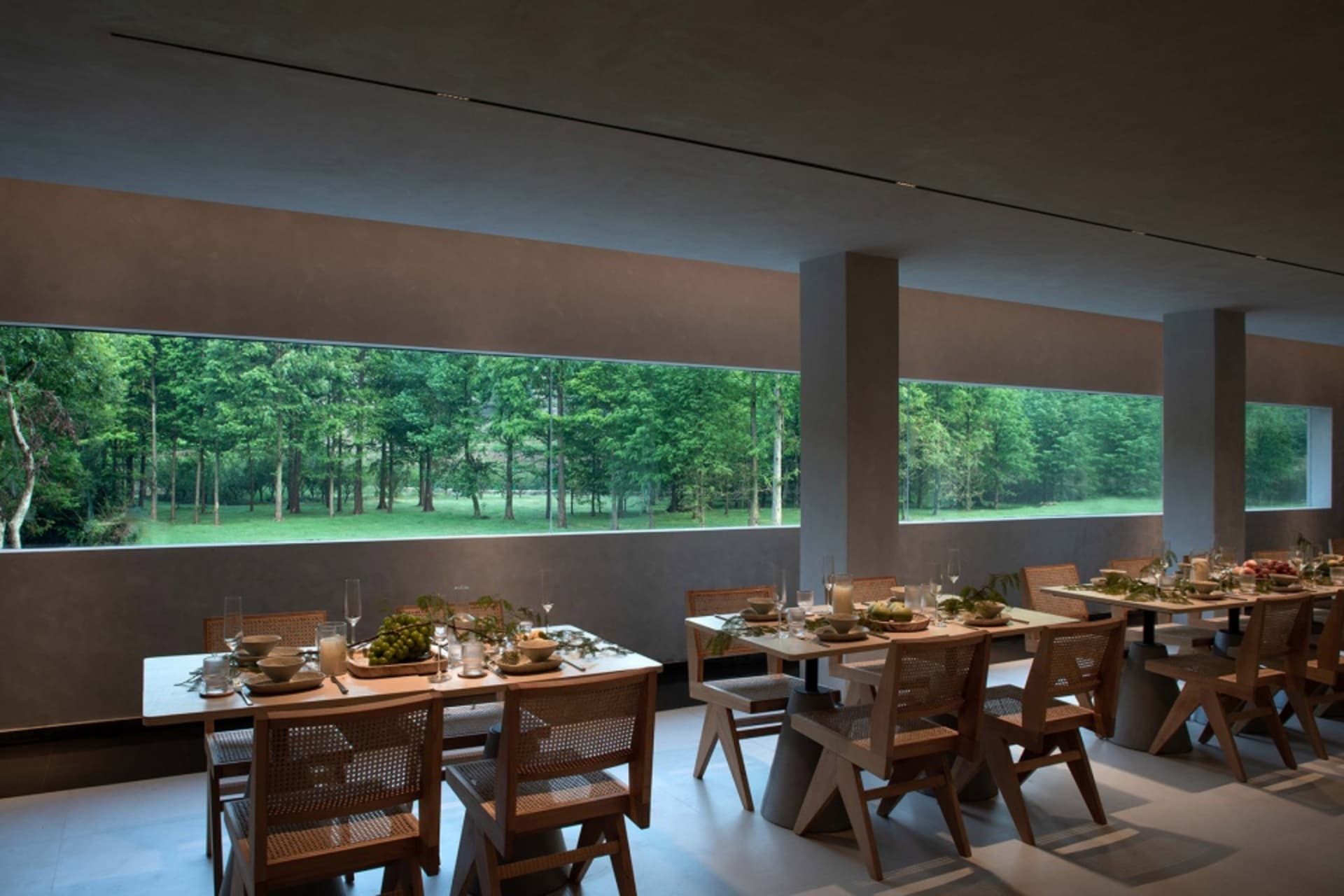 An industrial dining room with wooden tables, chairs, and a large glass window overlooking greenery.