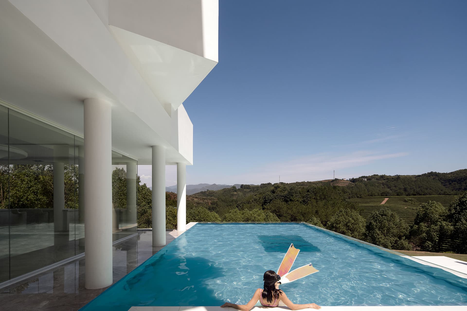 A minimalist residential home with a glass infinity pool overlooking scenic green hills under a clear blue sky.