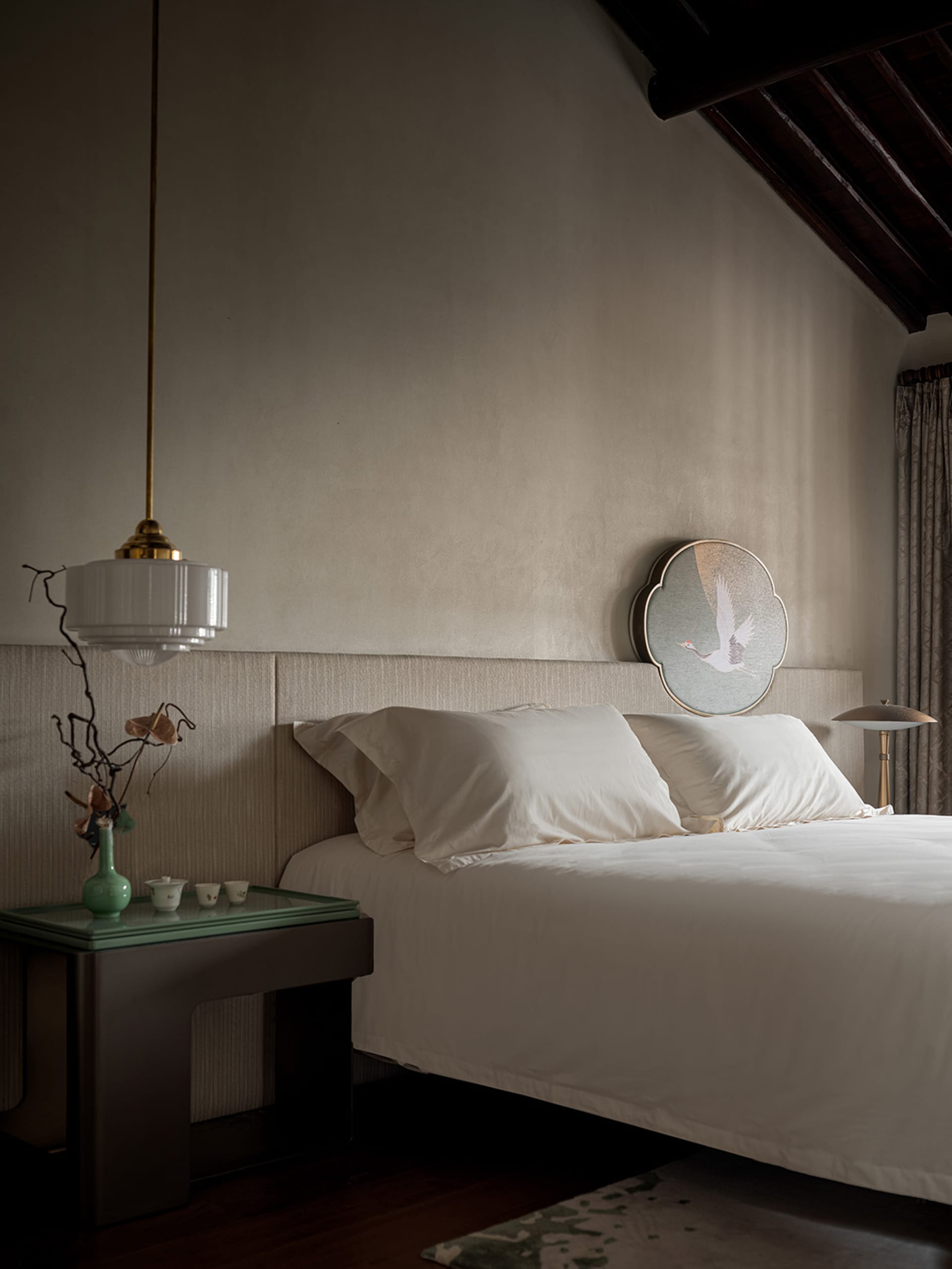 A minimalist bedroom featuring a white bed with a simple headboard, a side table with a vase and tea set, and a pendant light