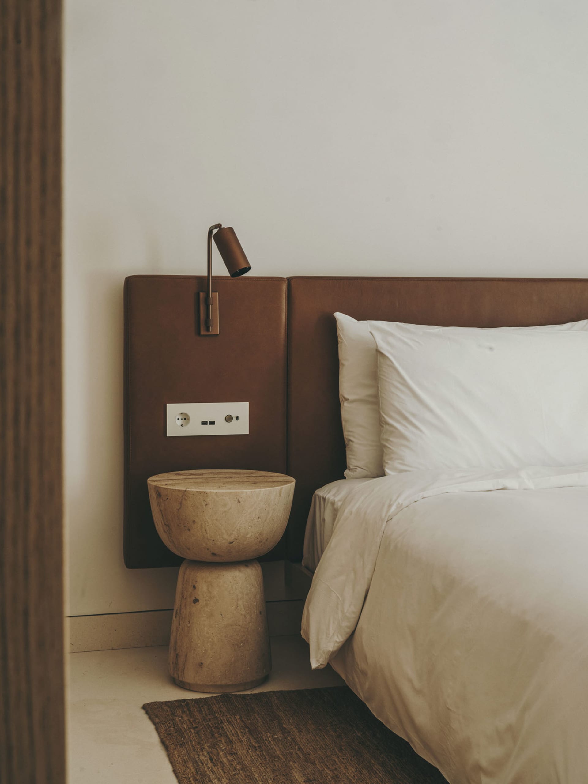 Modern minimalist bedroom featuring a bed with a brown leather headboard, white bedding, and a natural stone side table with 