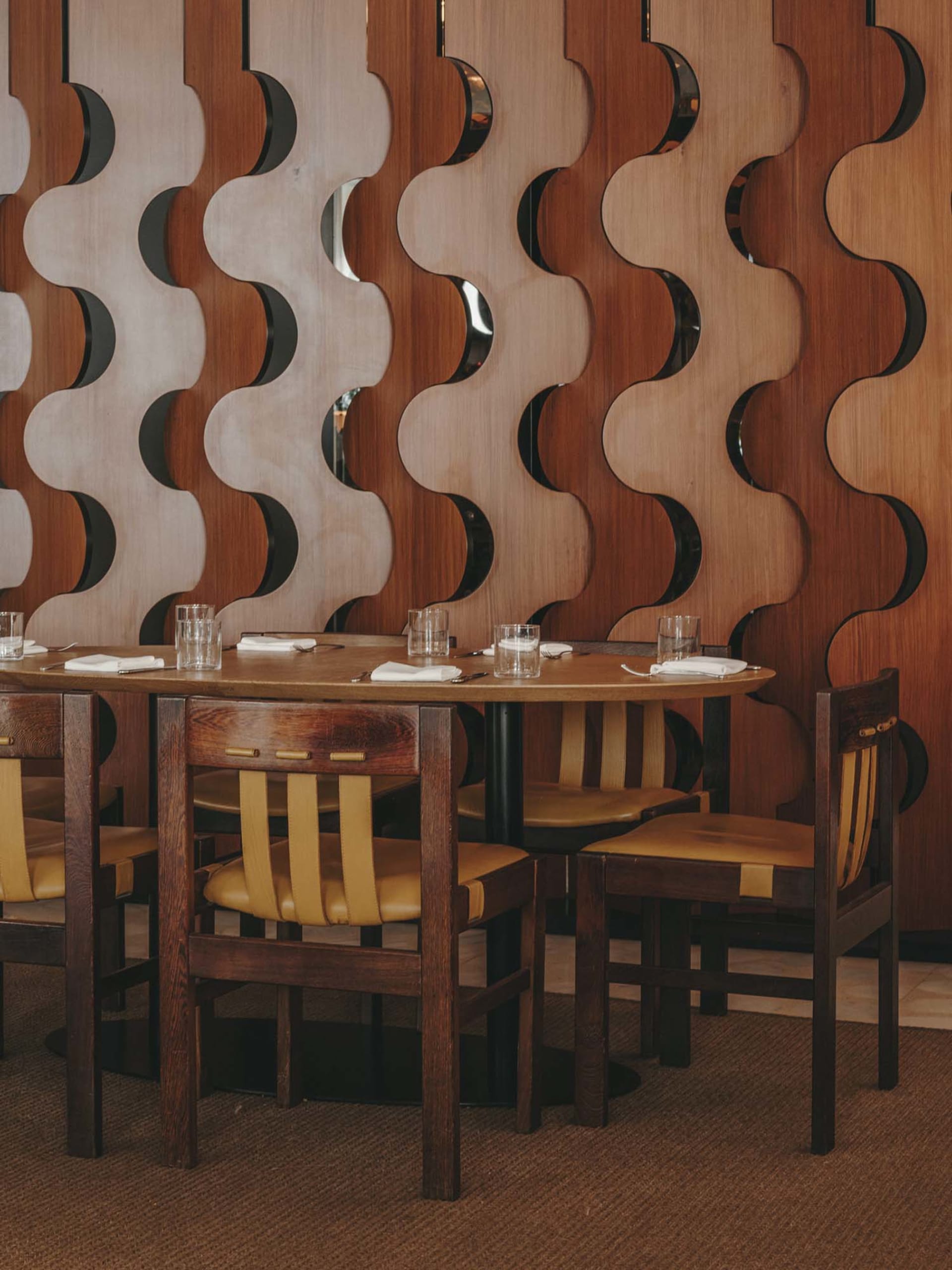 A modern restaurant interior with a wooden design featuring a round table, yellow chairs, and a decorative wave-patterned wal