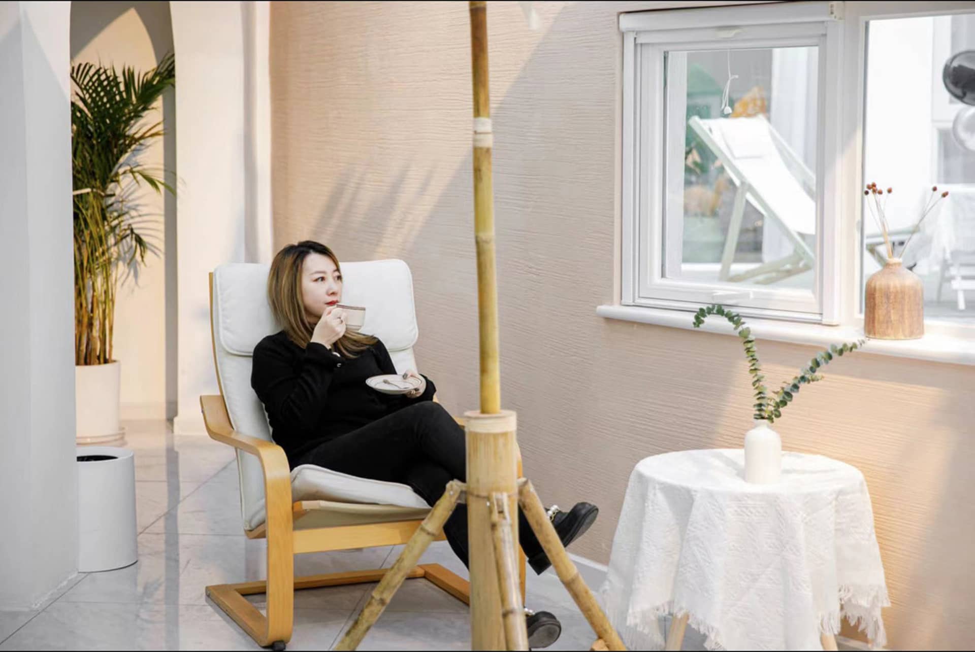 A woman sits in a minimalist relaxation space, enjoying a cup of tea in a cozy chair with a white cushion.
