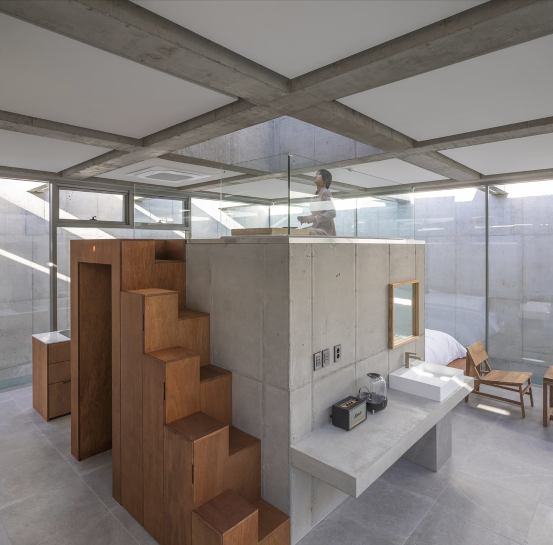 A minimalist living room with concrete walls, wooden stairs, and a glass partition.