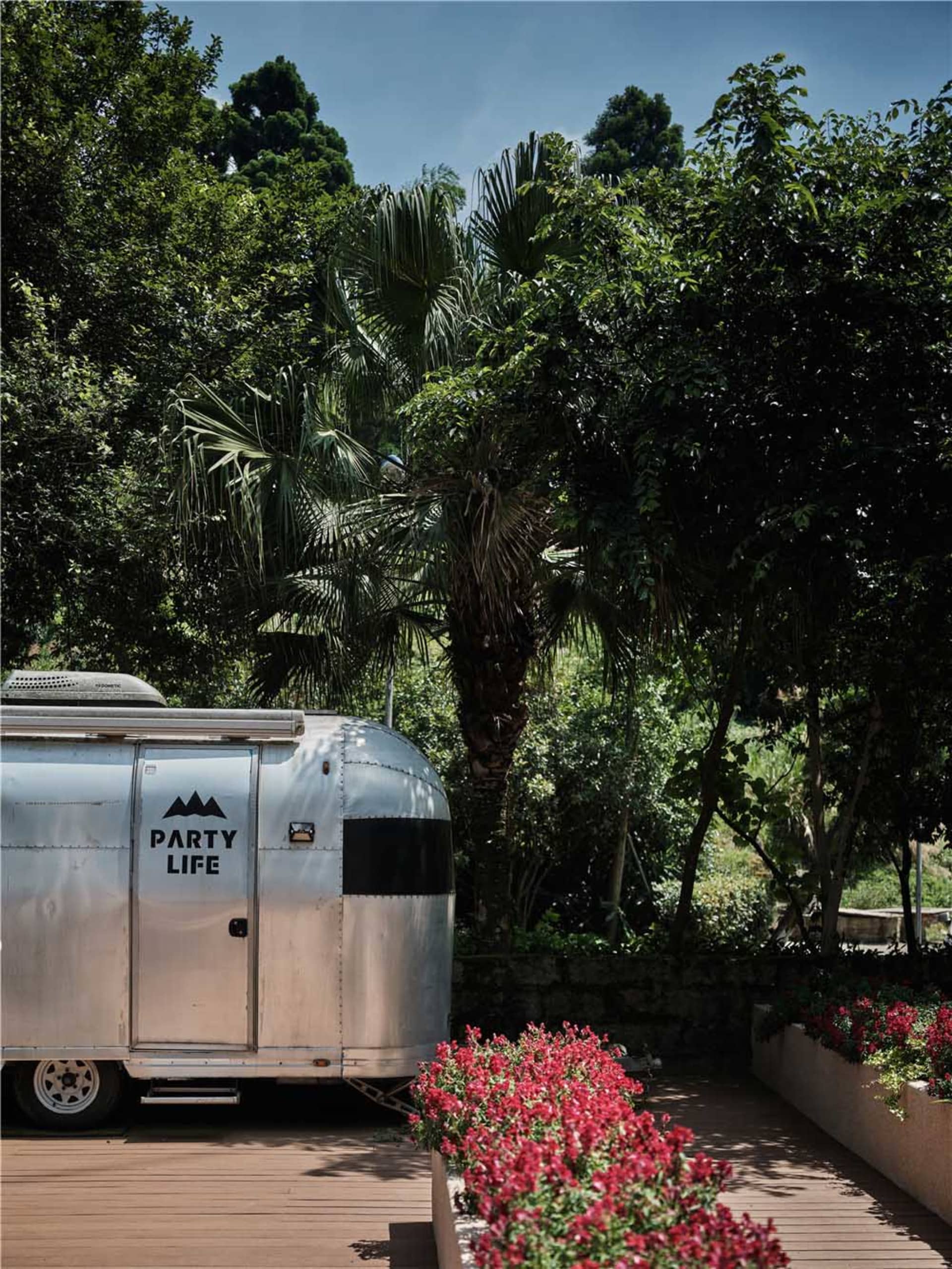 A silver, metallic camping trailer with the text 'PARTY LIFE' on its side, parked on a wooden deck surrounded by lush greener