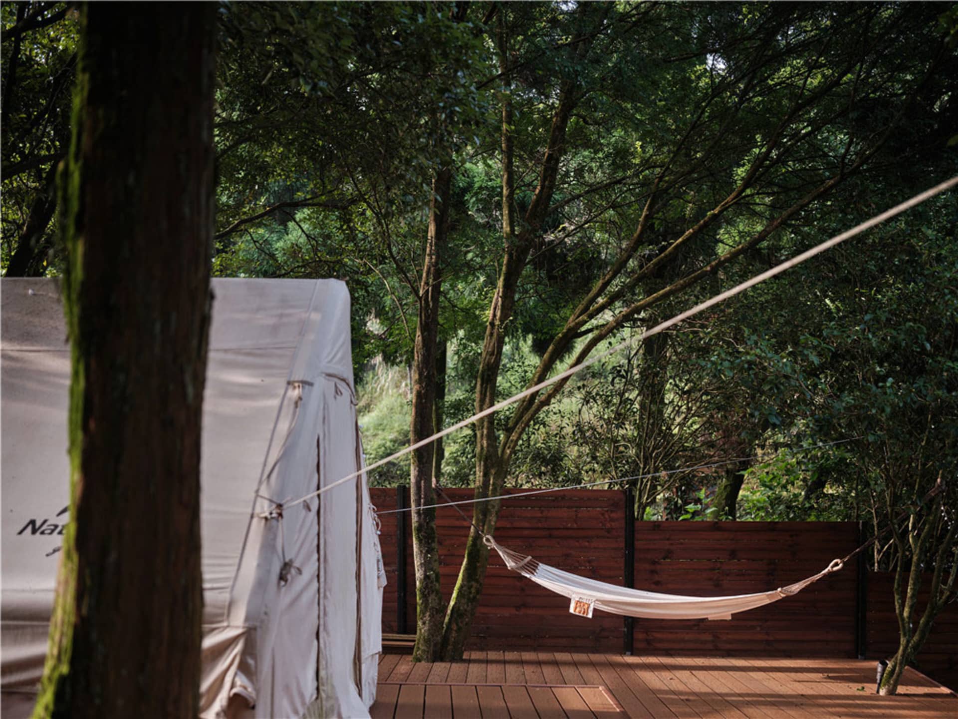 A white hammock hanging from a wooden frame on a deck surrounded by trees and foliage.