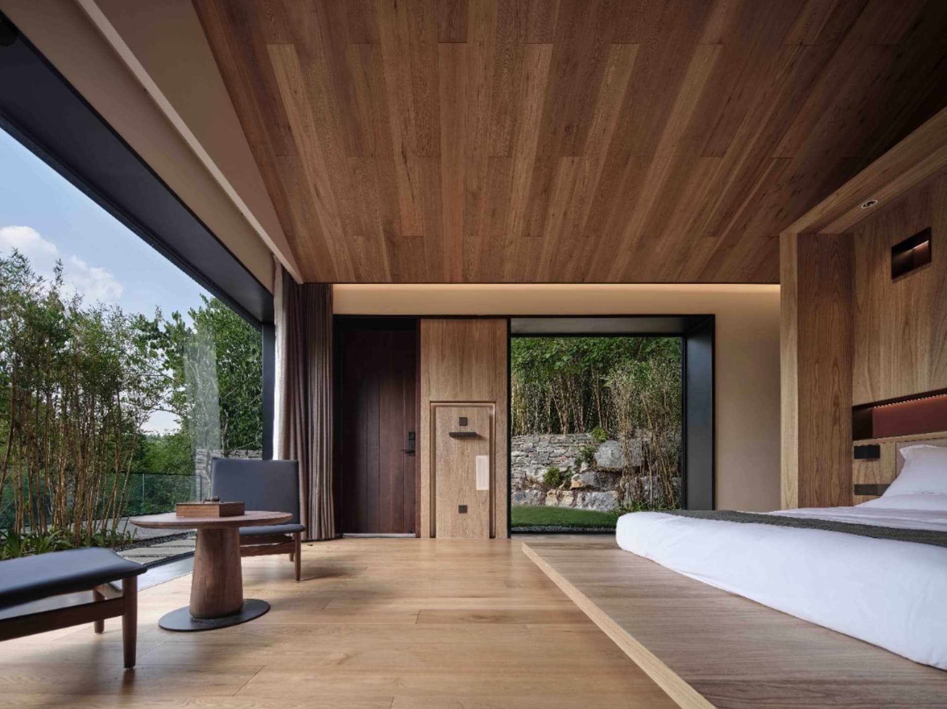 A modern bedroom with wooden walls and a large window overlooking greenery.