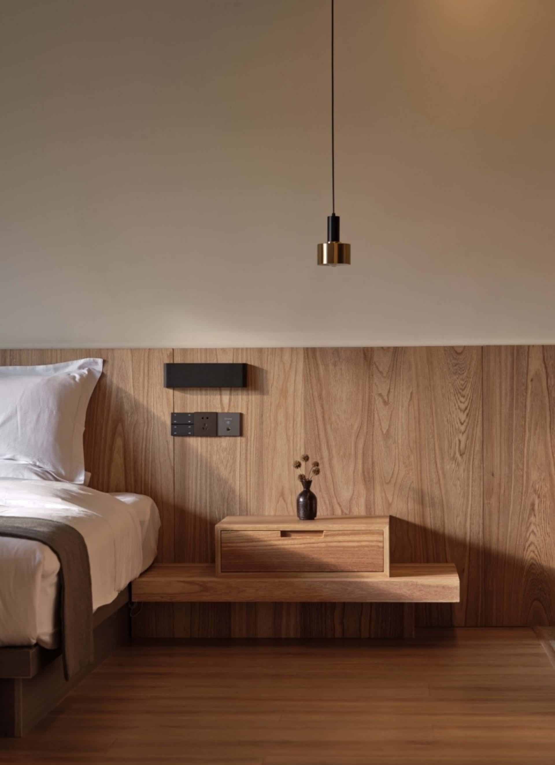 A minimalist bedroom featuring a wooden bed, wooden headboard, and wooden floating nightstand with a vase and a lamp hanging