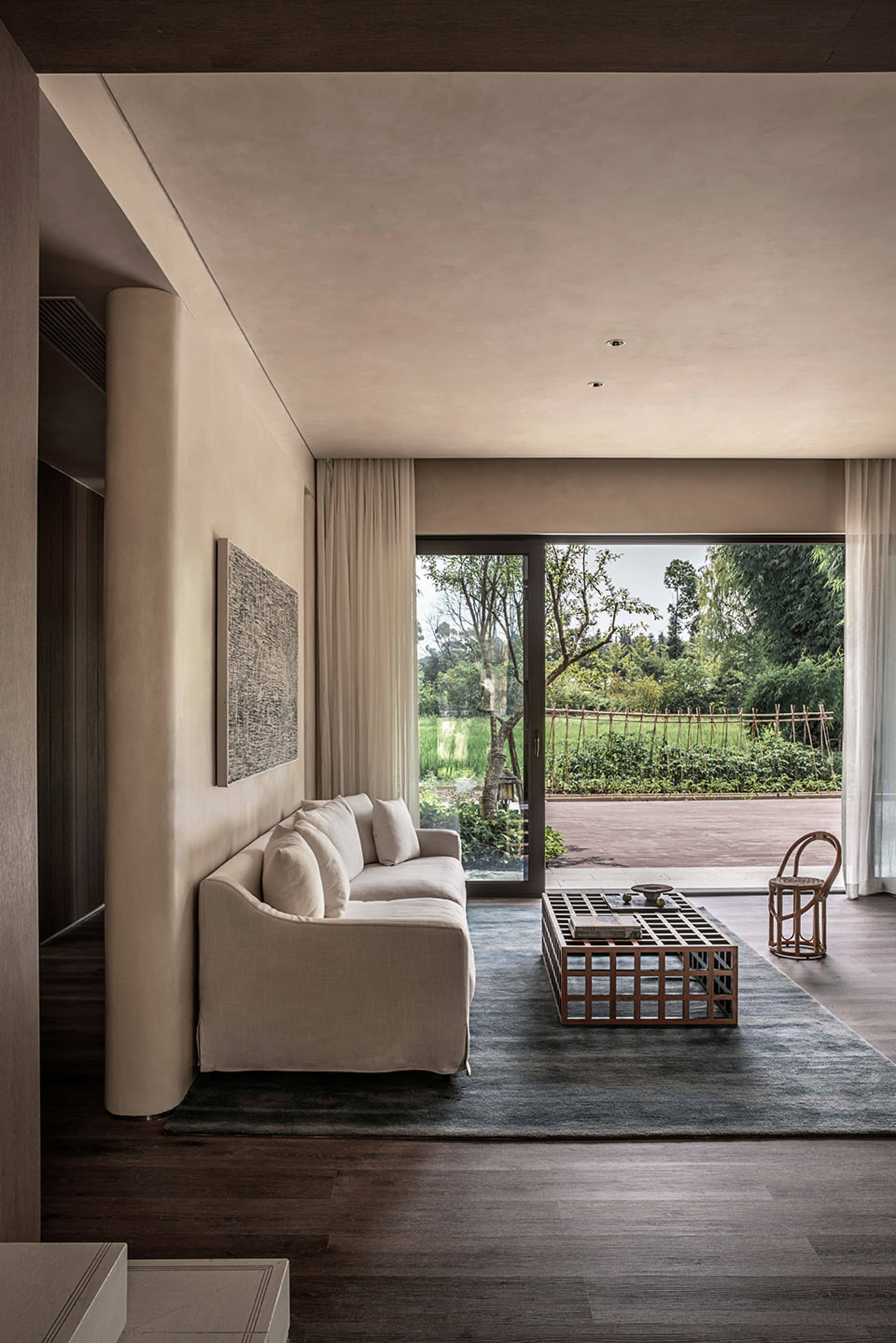 A modern living room with a beige sofa, a wooden coffee table, and a large window overlooking a green outdoor area.