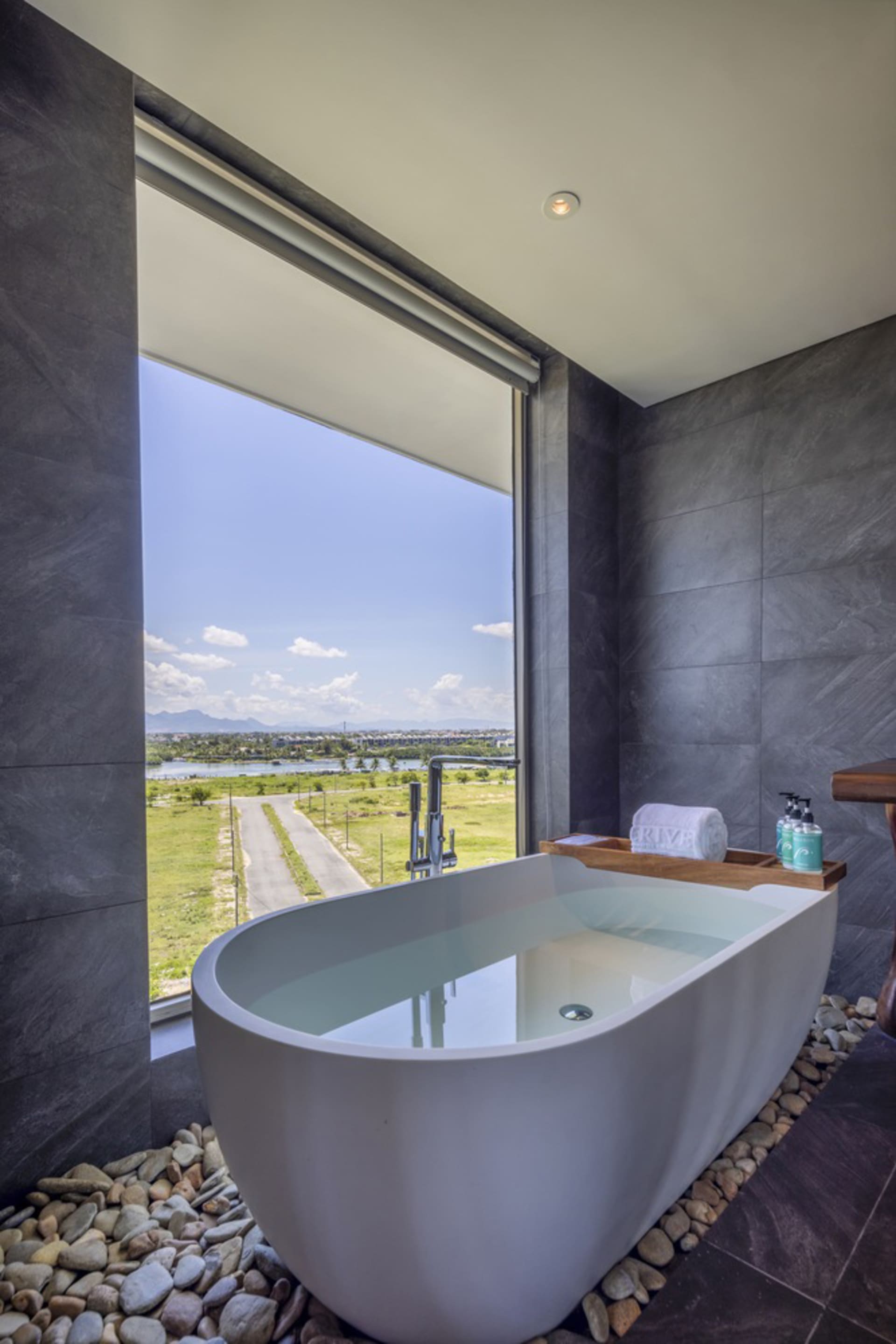 A modern bathroom with a white freestanding bathtub surrounded by rocks, featuring a large window overlooking a peaceful land