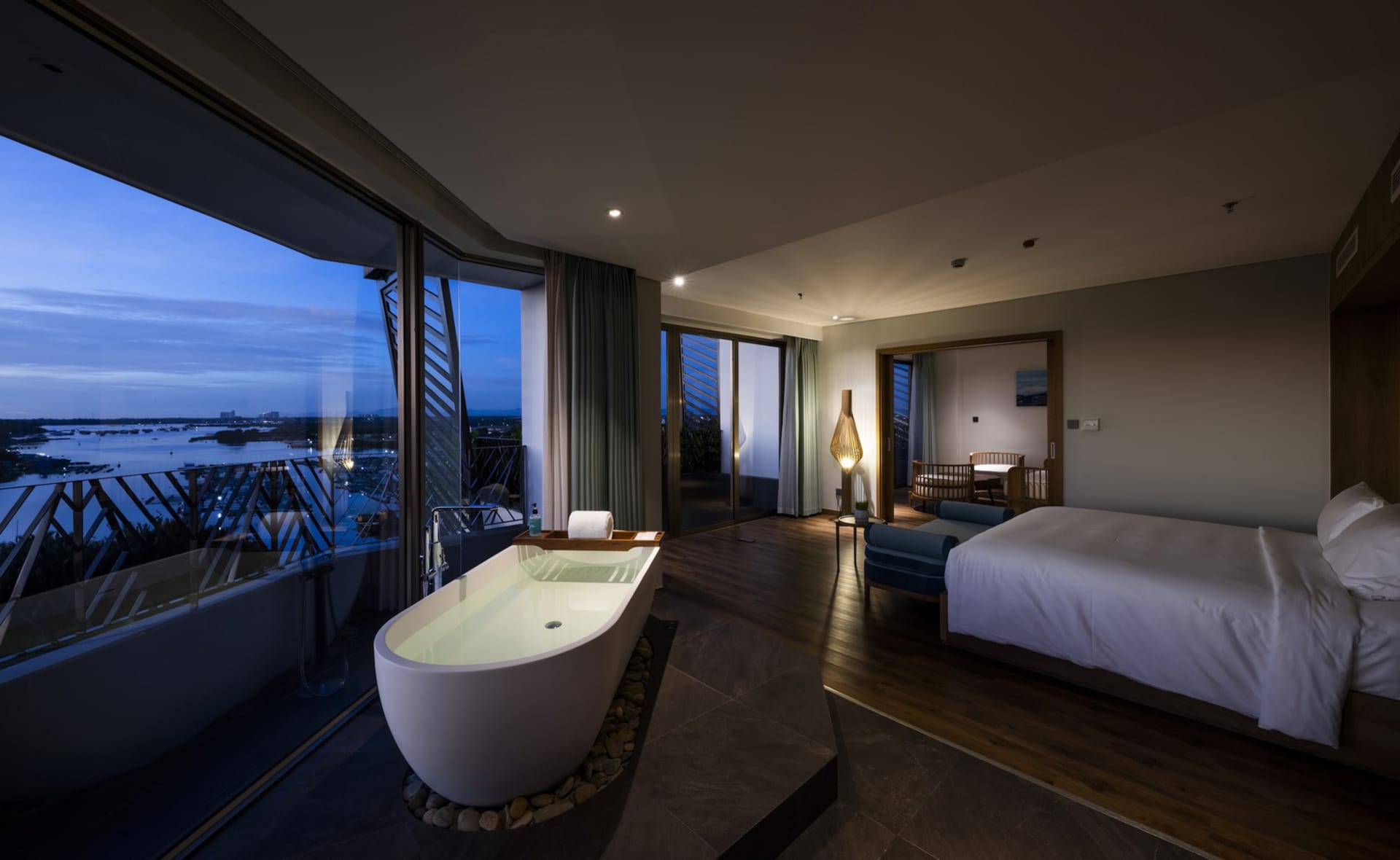 A modern bedroom with a white bed, wooden floor, and large windows overlooking a cityscape at dusk.