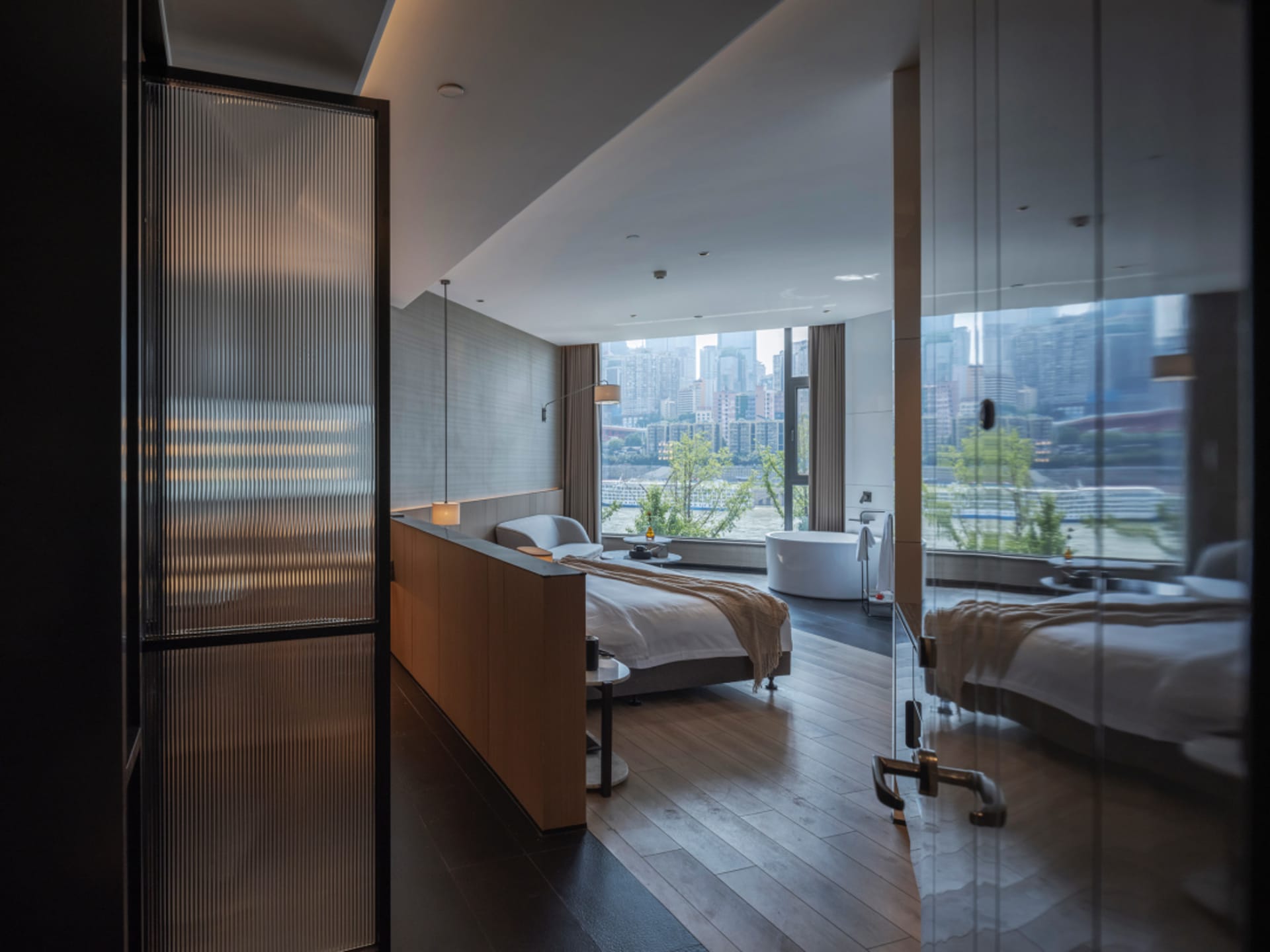 A modern minimalist bedroom with wooden flooring and a sleek, white bathtub overlooking a cityscape.
