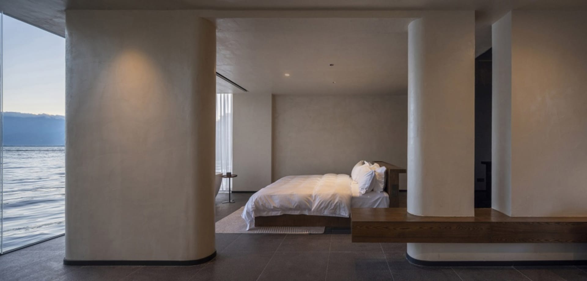 A minimalist bedroom with concrete walls and floor, featuring a bed with white bedding and wooden accents.
