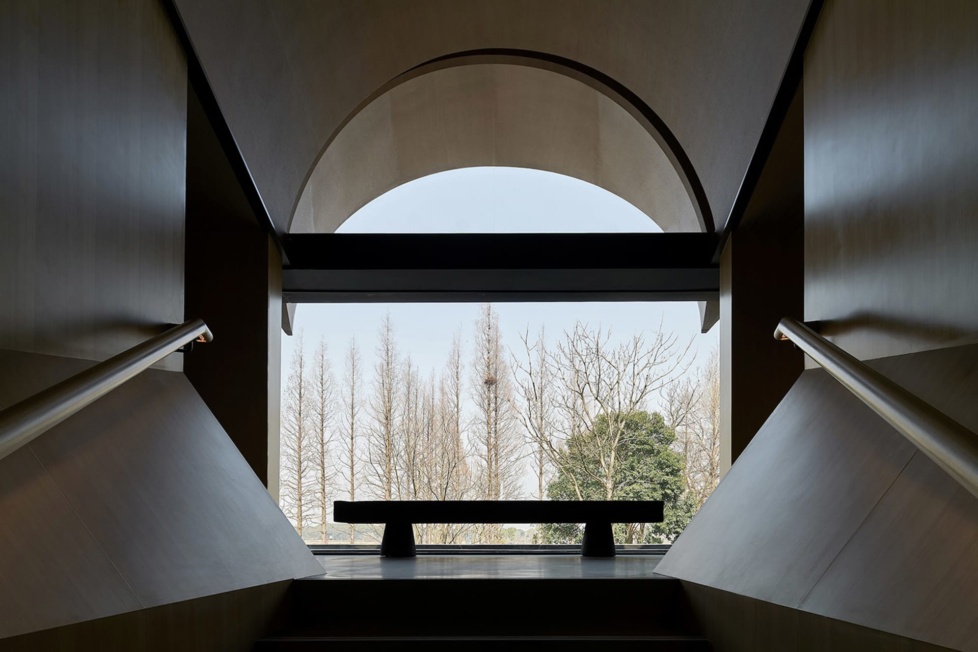 Concrete staircase viewed from above, featuring an arched window with a bench and a scenic outdoor view of bare trees.