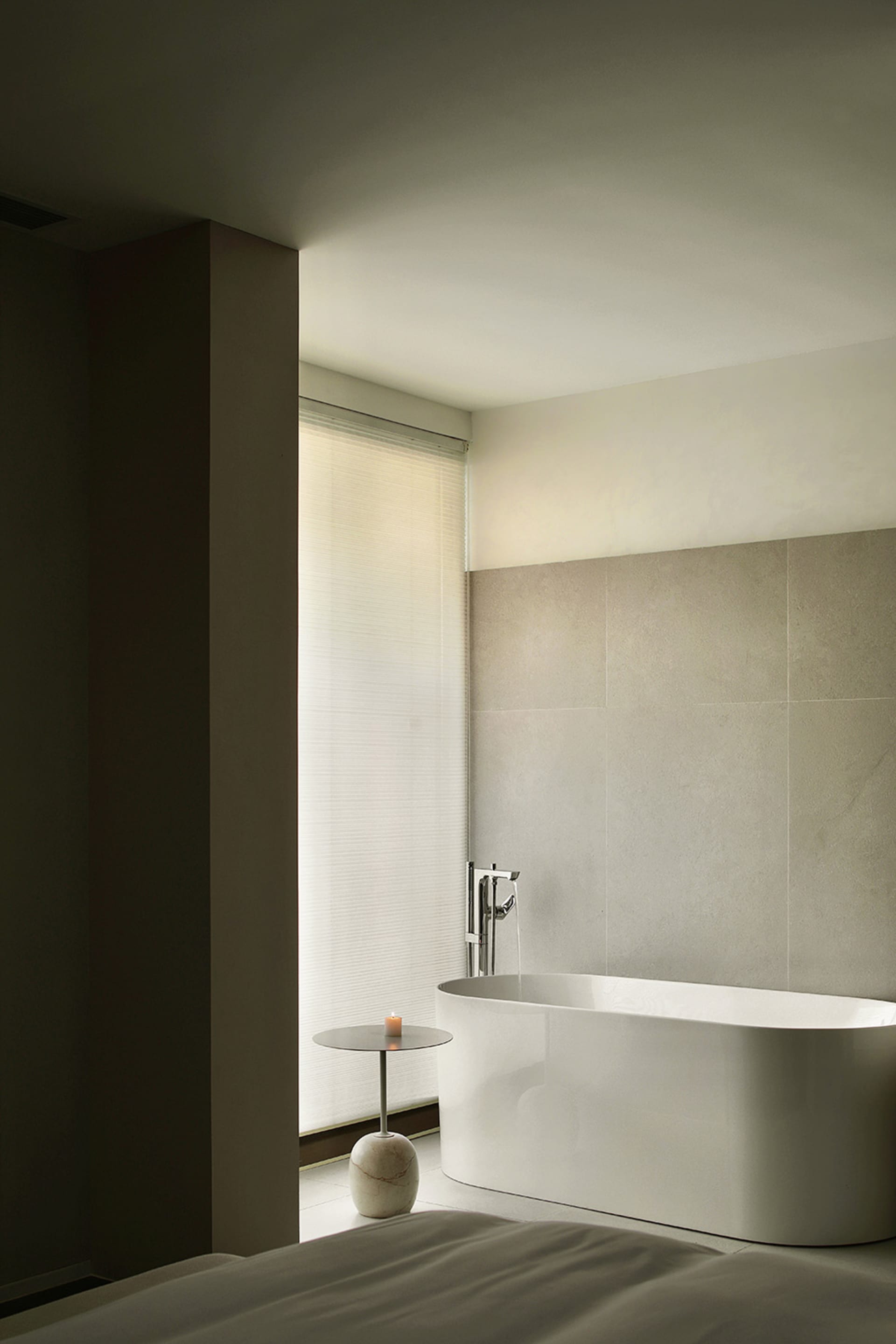 A minimalist bathroom with a white ceramic bathtub, a small marble table with a candle, and a window with blinds.