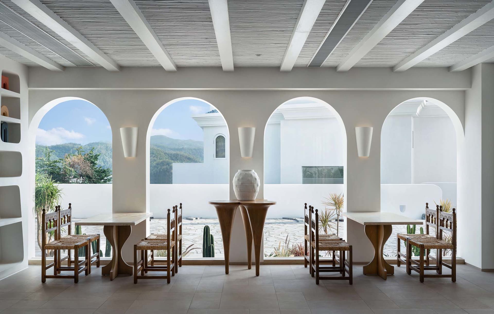 A minimalist restaurant interior with wooden tables and chairs, open windows, and a view of the mountains.
