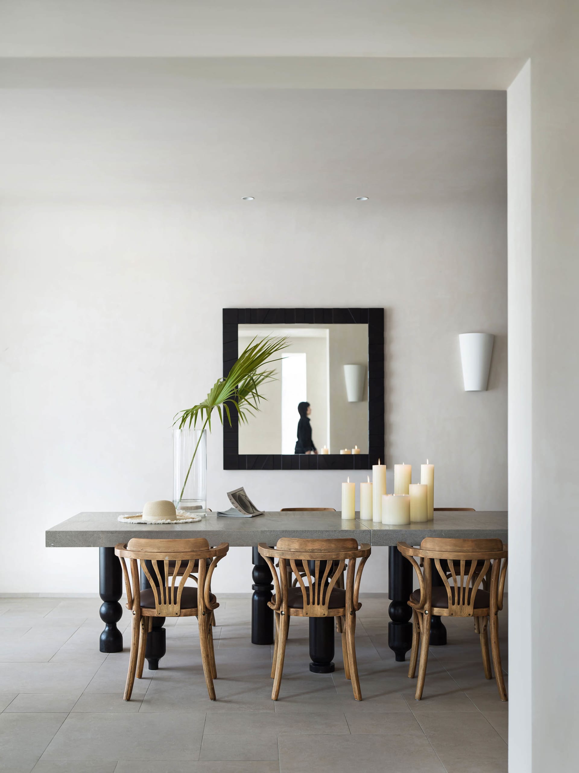 A modern minimalist dining room featuring a concrete table and wooden chairs, with a large mirror on the wall, candles, and a