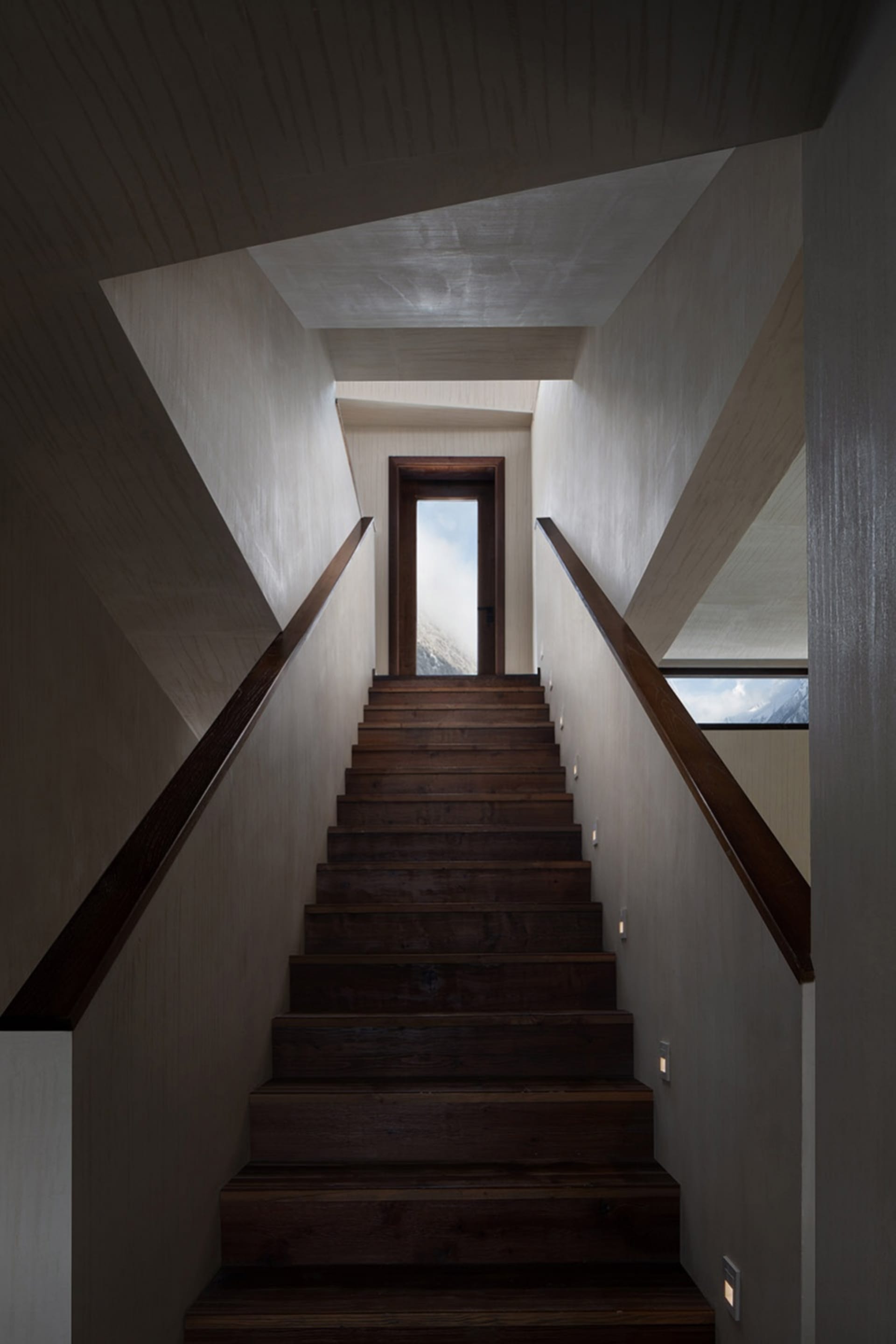 Modern, wooden staircase with industrial design elements, featuring a hallway with a door opening towards a sky view.