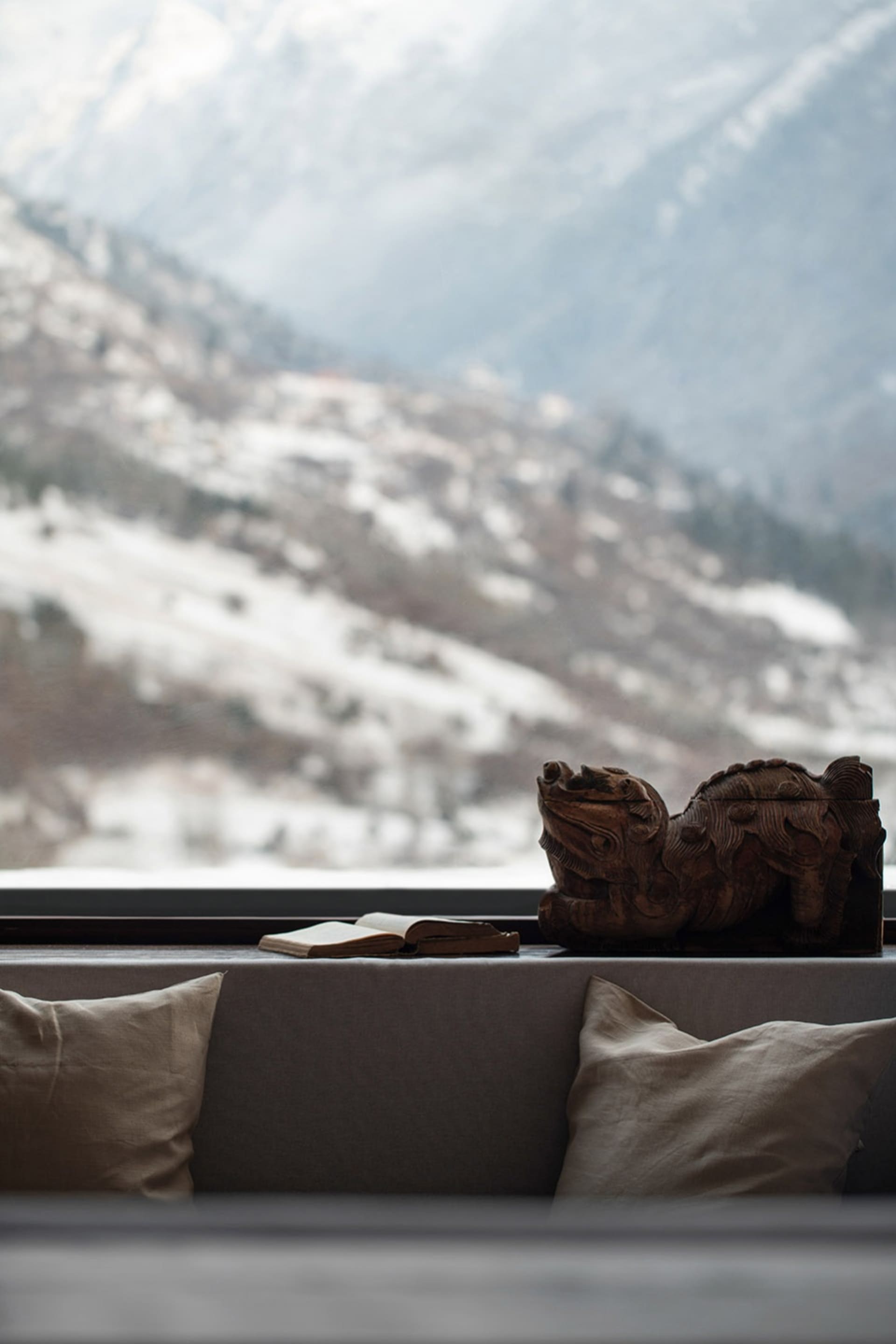 A cozy living room with a carved wooden lion statue, a book, and cushions overlooking a snowy mountain landscape.