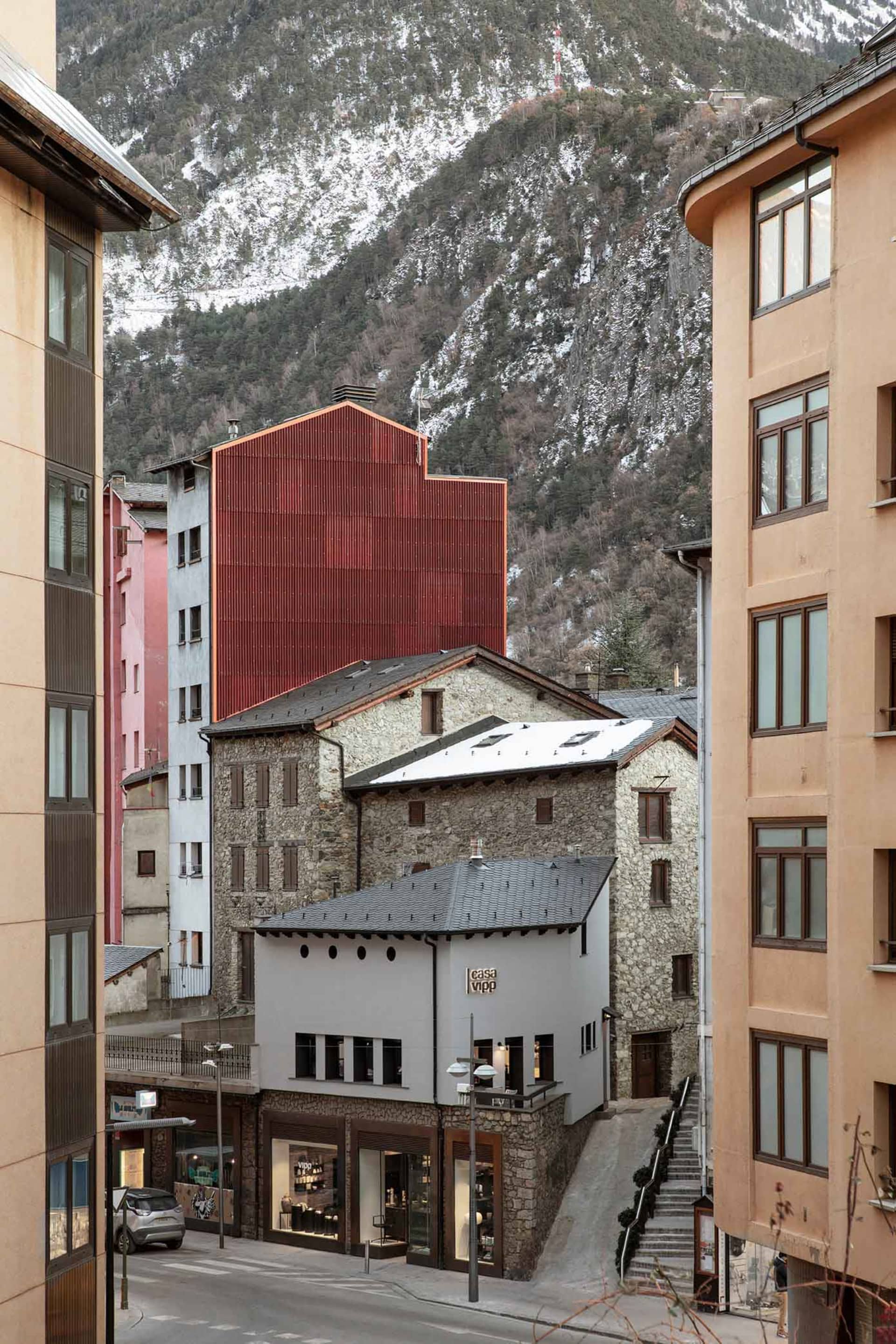 Stone and concrete building in a narrow street with a modern red structure in the background.
