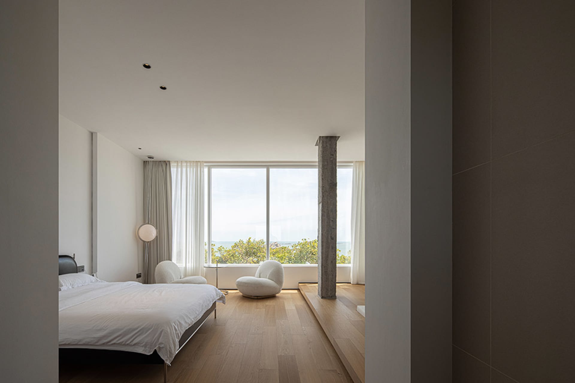A modern bedroom with a white bed, white chairs, and wooden flooring, featuring a large window with curtains and a concrete c