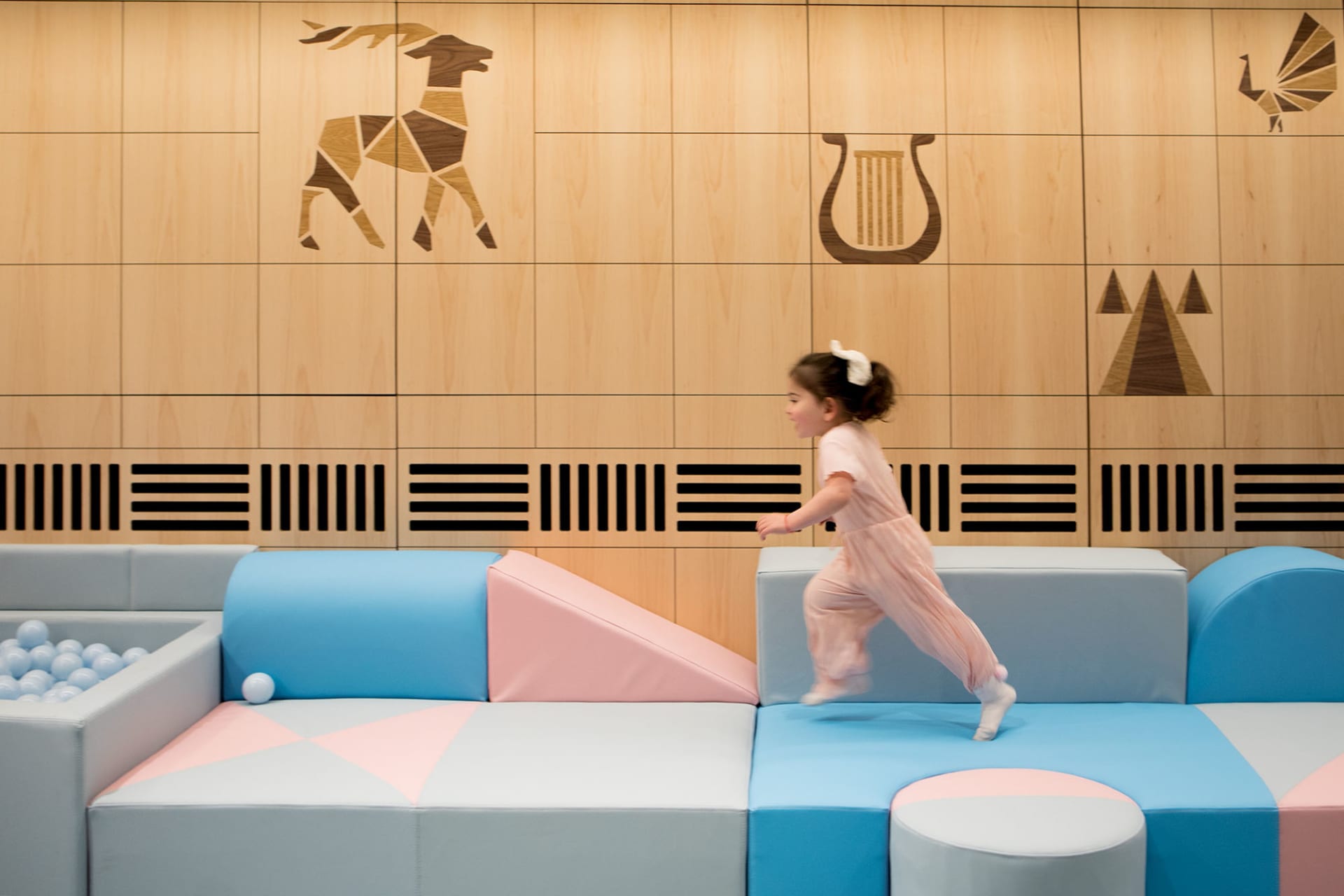 A child in a pink outfit running on soft play structures in a modern playroom