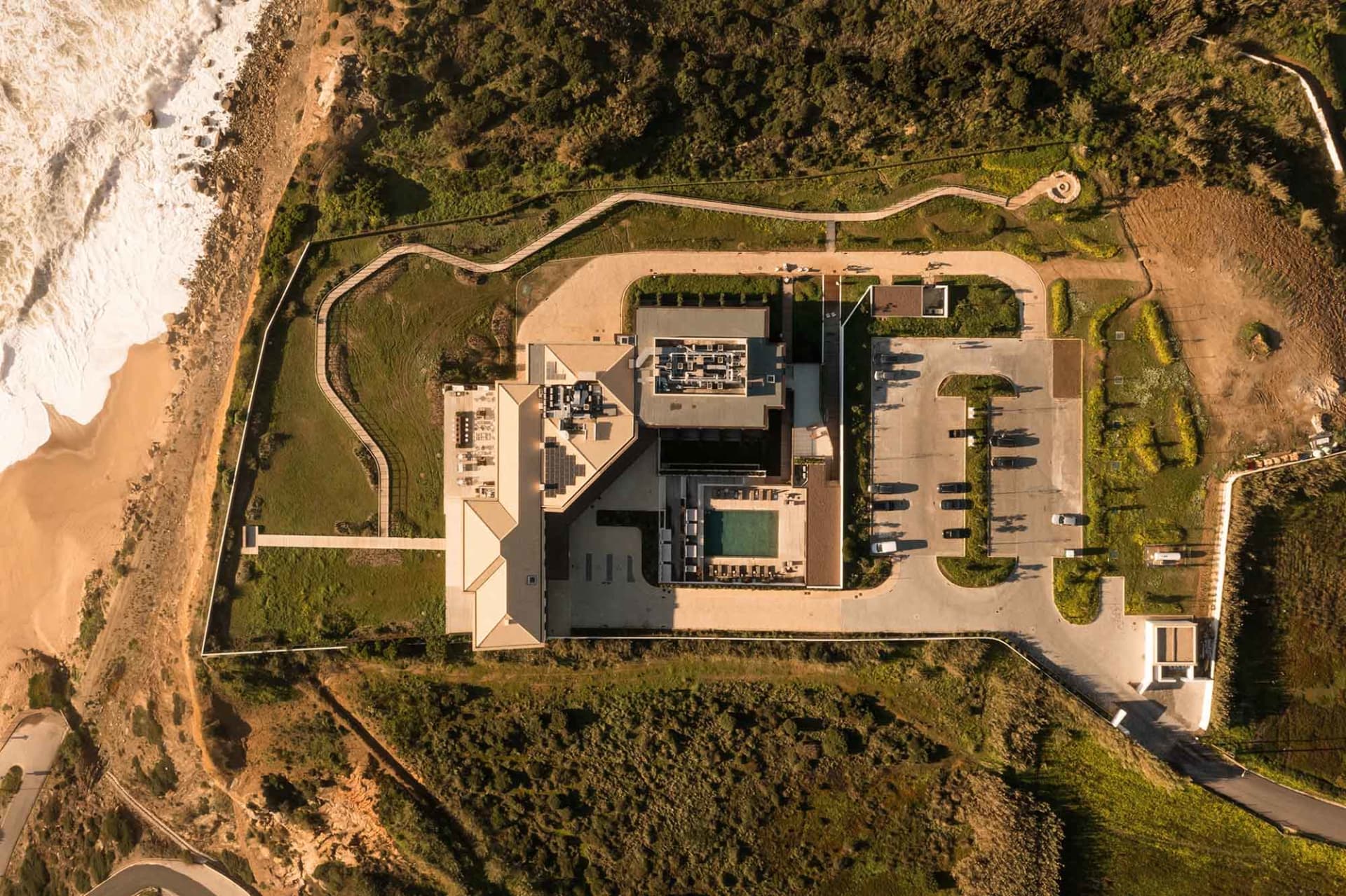 Aerial view of a modern residential building with a rooftop pool, surrounded by greenery and a beach.
